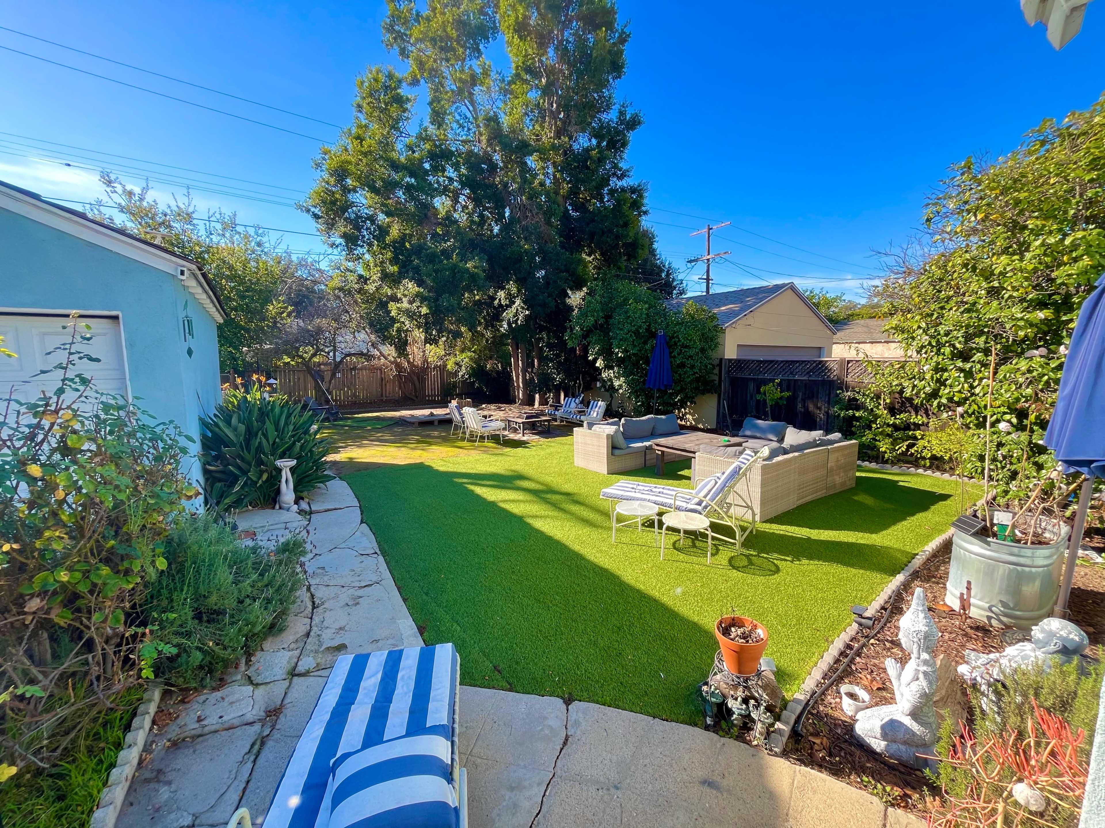 The image shows a backyard with artificial grass, outdoor seating areas, and surrounding greenery.