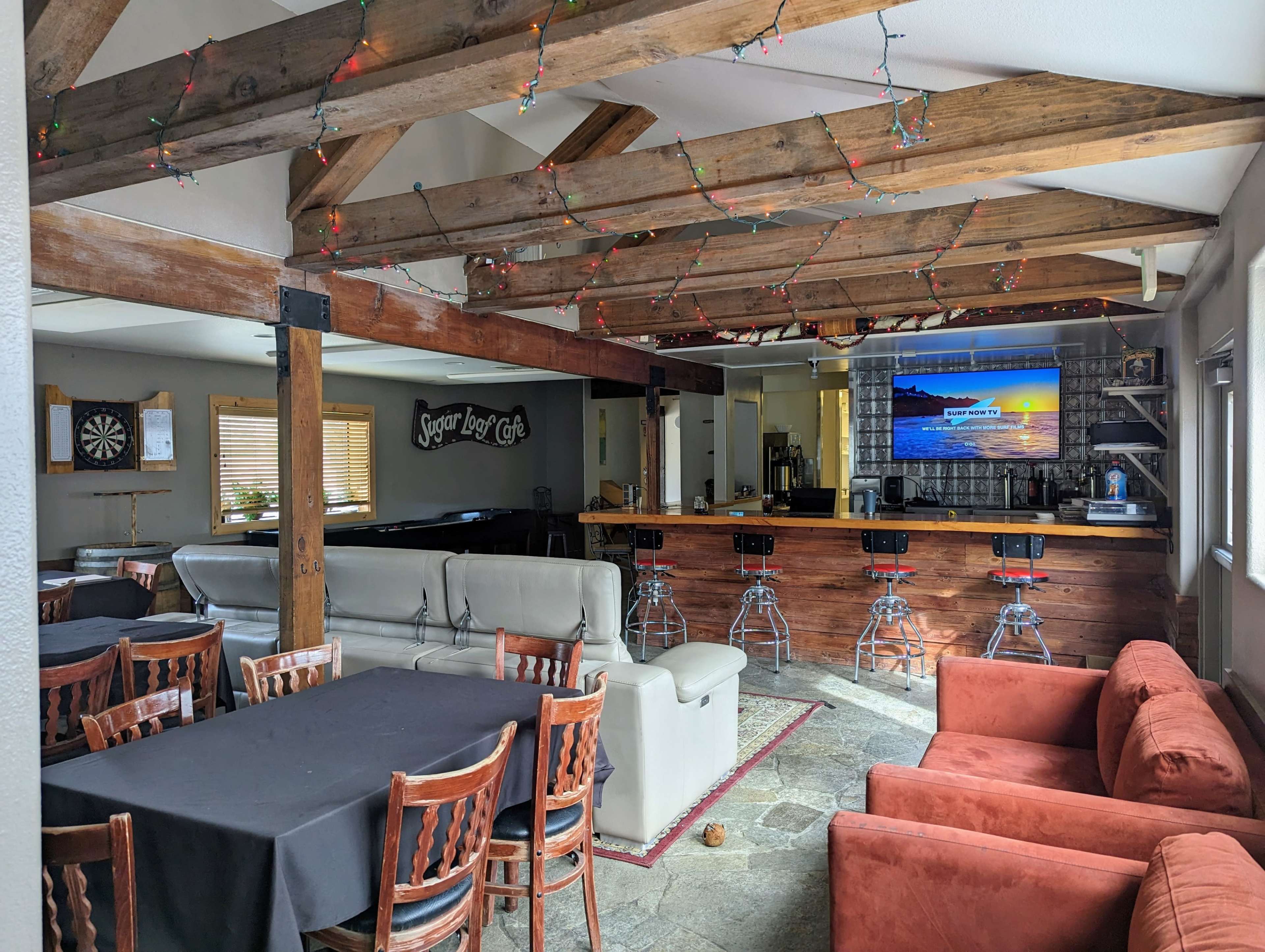 A cozy recreational room with wooden beams, a bar counter, seating areas, and a large television displaying a sunset scene.