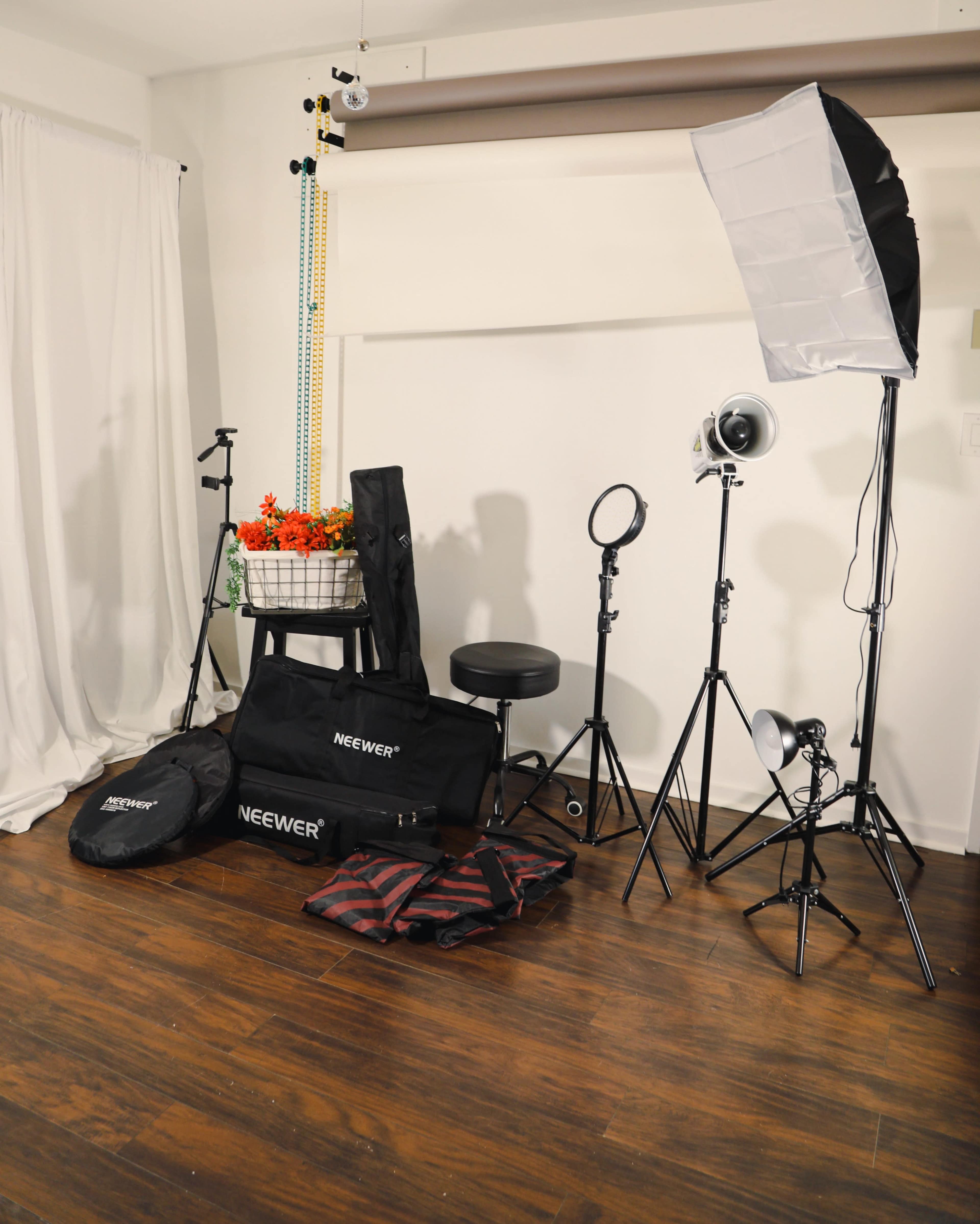 A photography setup featuring various lighting equipment, bags, and a stool arranged in a corner of a room with a white backdrop.