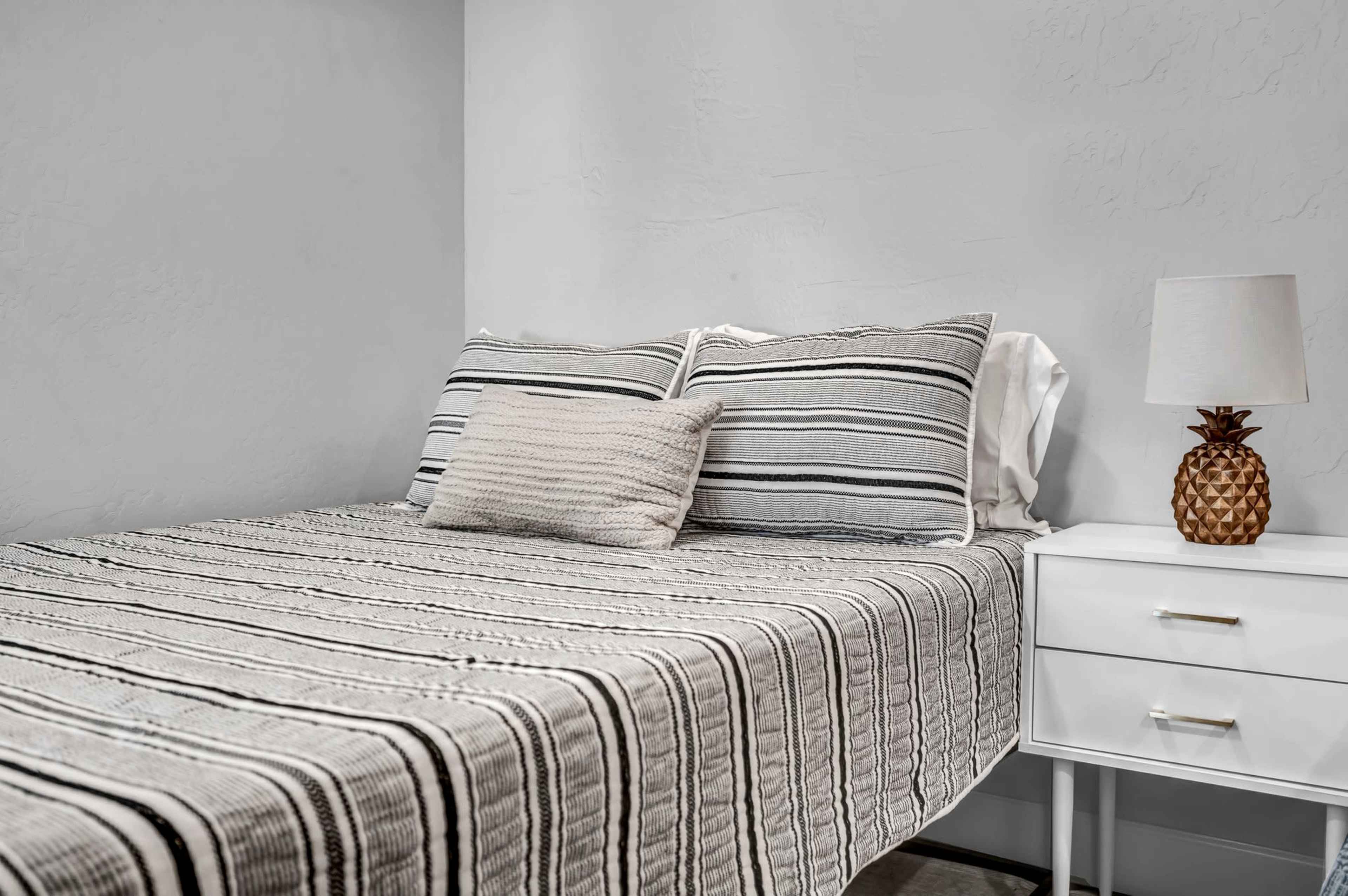 A striped bedspread covers a bed with two pillows and a decorative throw pillow, accompanied by a bedside table with a lamp shaped like a pineapple.