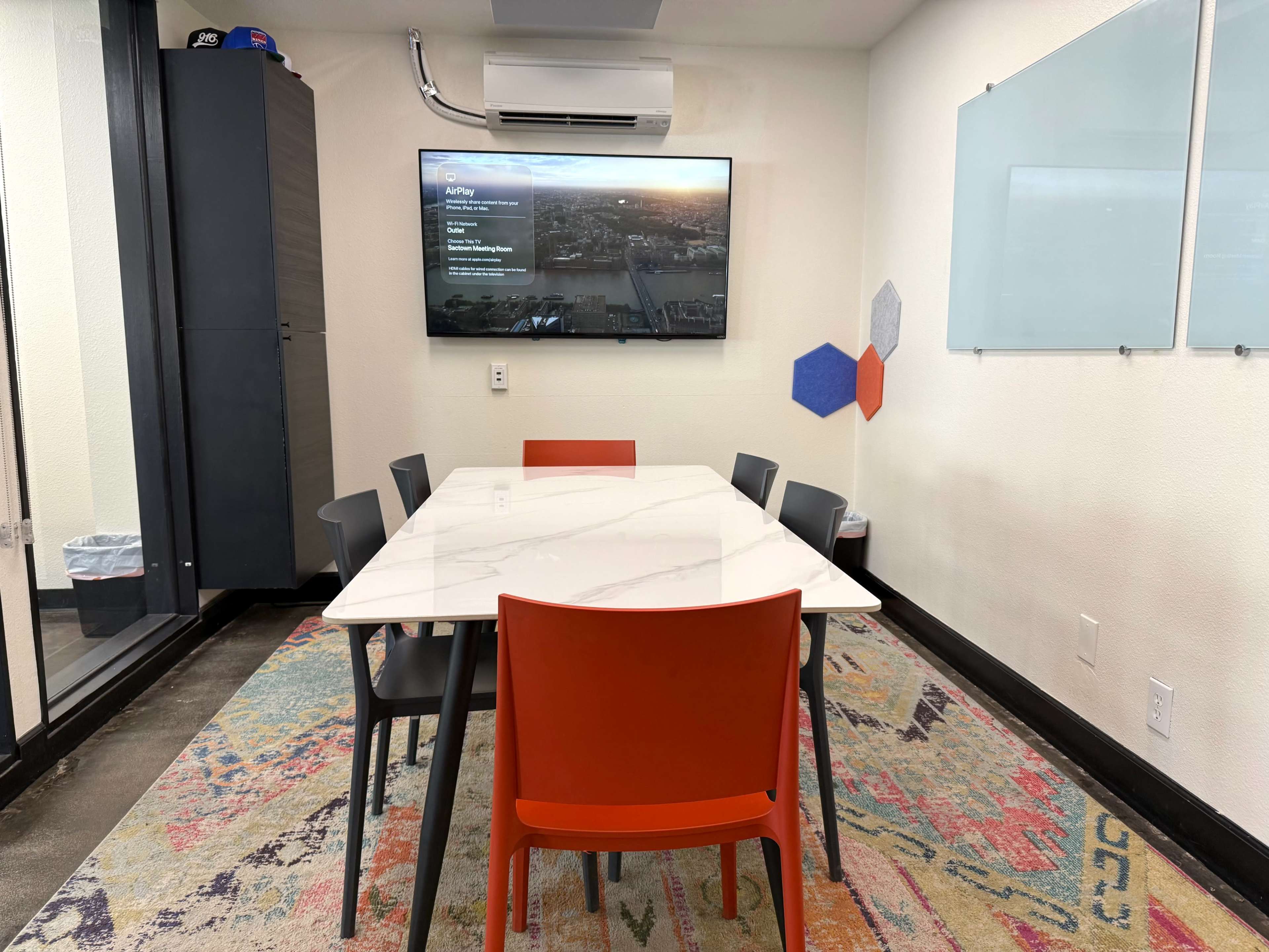 A modern meeting room features a large white table surrounded by black and orange chairs, with a flat-screen TV mounted on the wall.