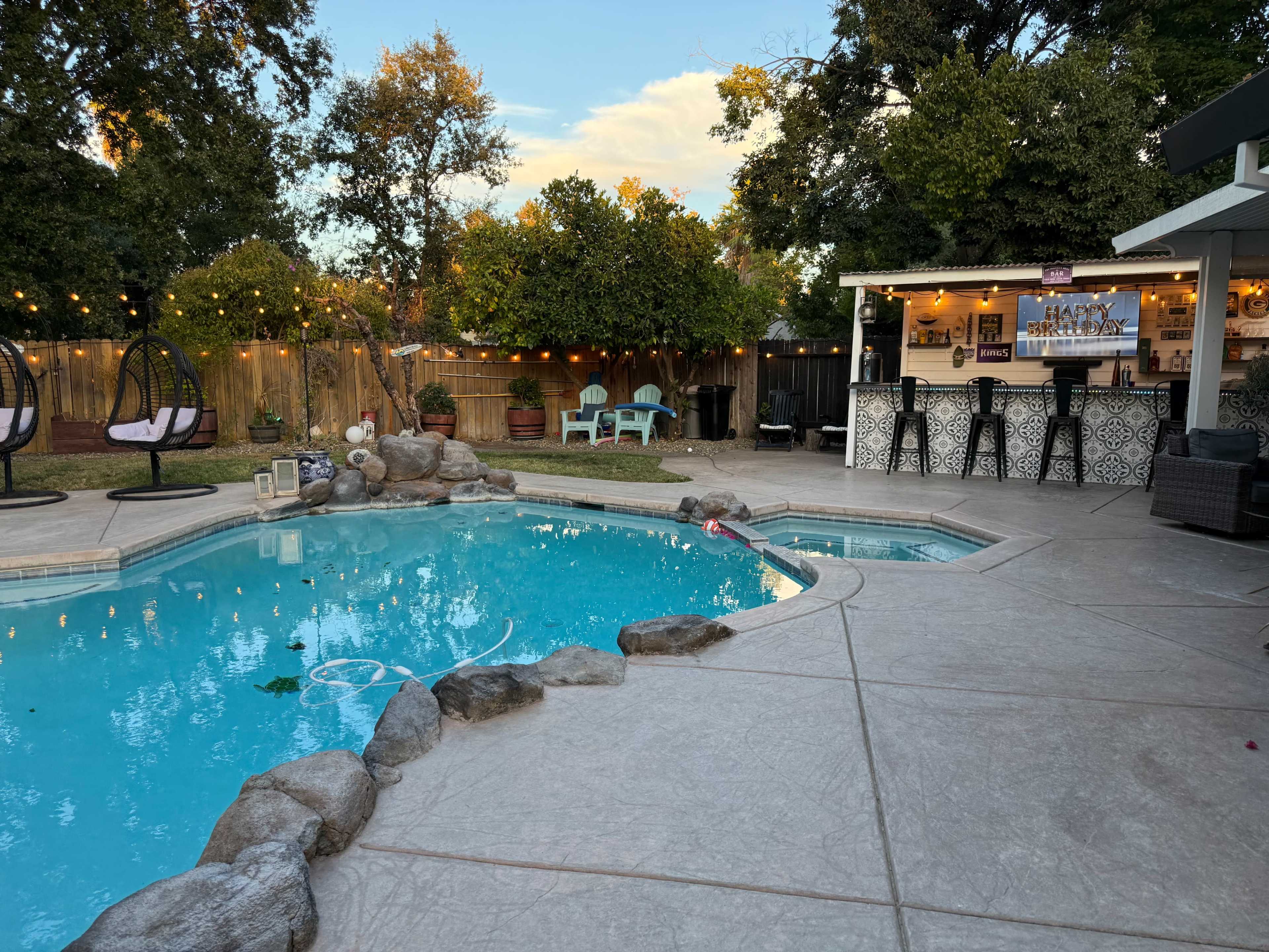 A clear, blue swimming pool is surrounded by stone detailing, with a patio area featuring hanging chairs and a bar in the background.