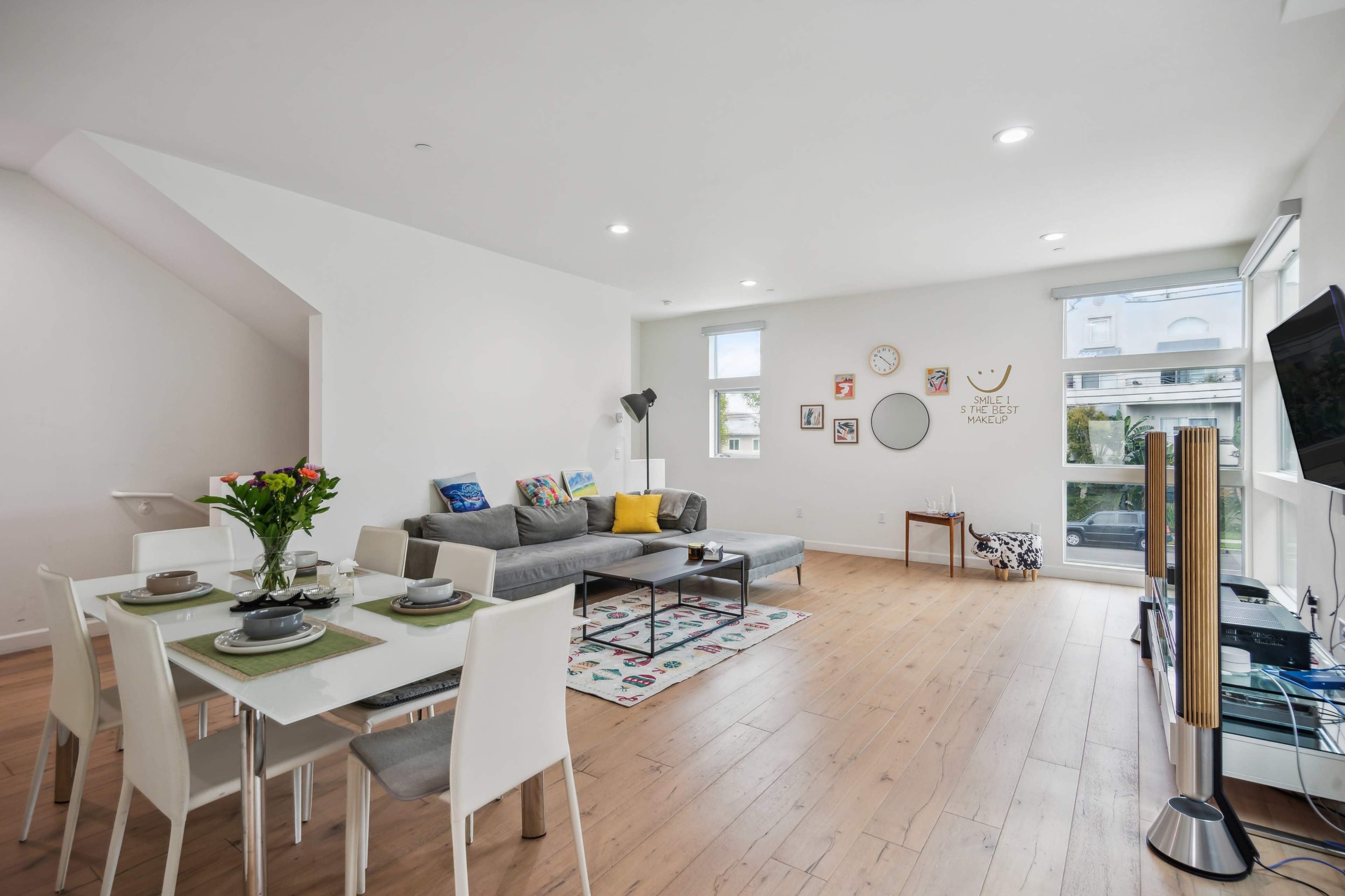 A modern, open living area with a dining table set for four, a sectional sofa, and large windows allowing natural light to enter.