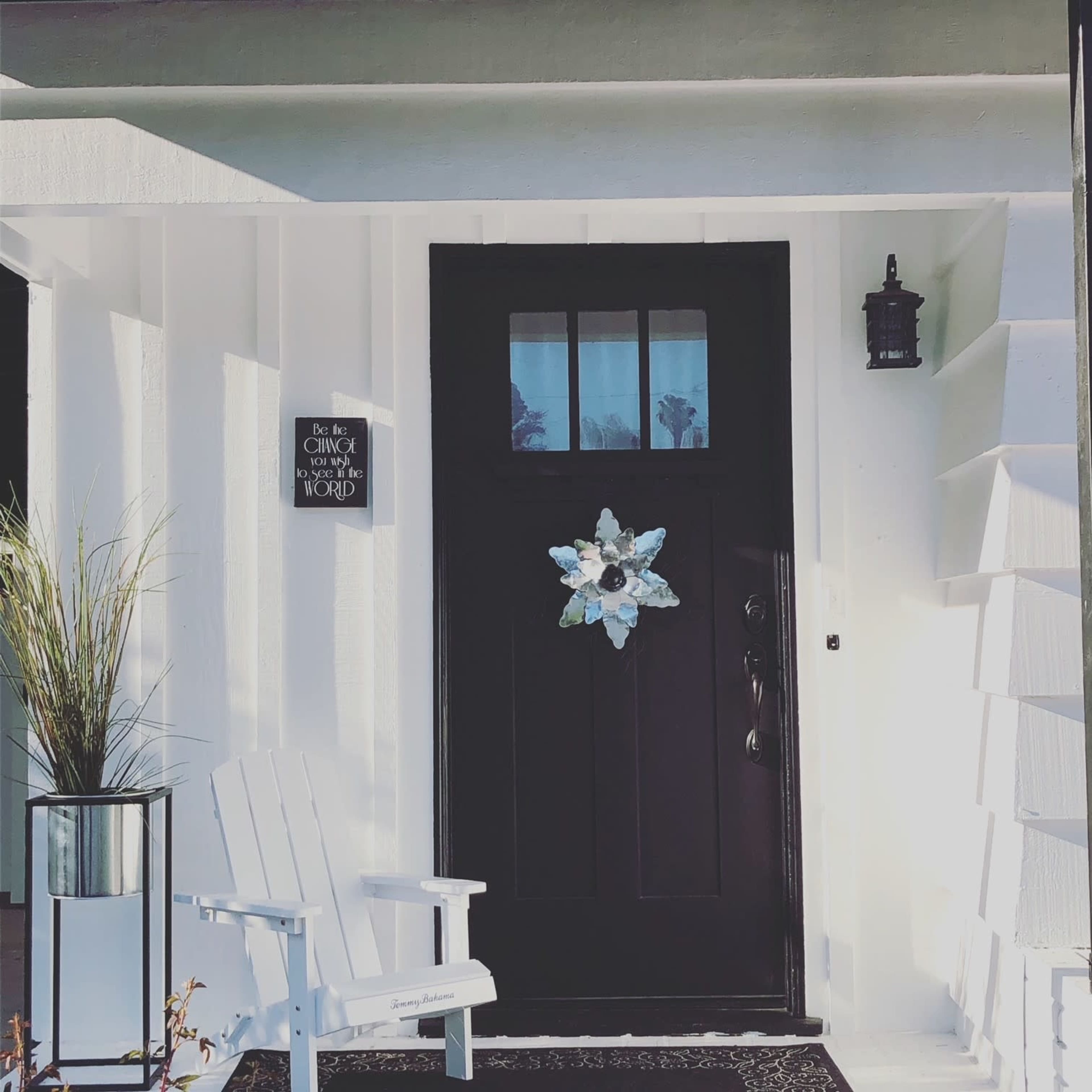 A black front door adorned with a decorative wreath, flanked by a white adirondack chair and a potted plant on a stand.