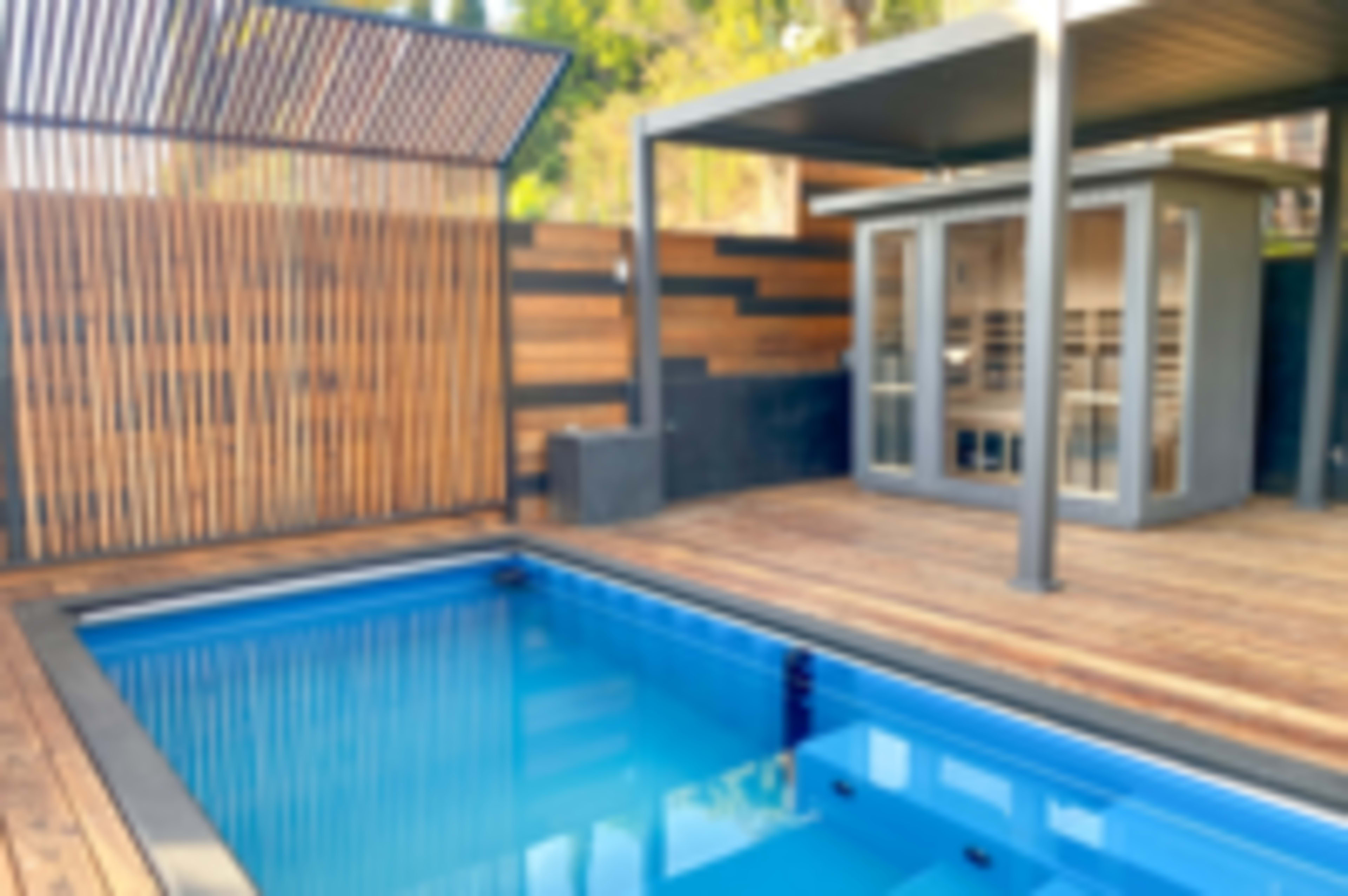 The image shows a modern outdoor space featuring a rectangular swimming pool beside a wooden deck and a sauna.