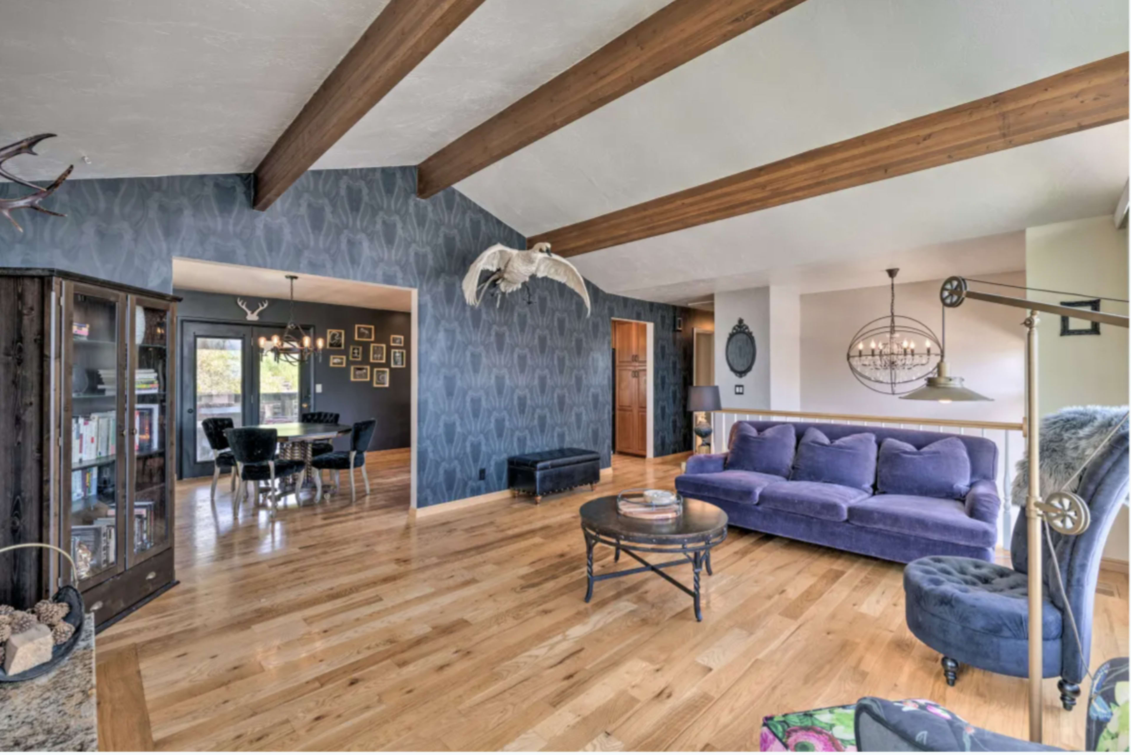 The spacious living room features hardwood floors, vaulted ceilings with exposed wooden beams, a purple sofa, and large windows that offer natural light.
