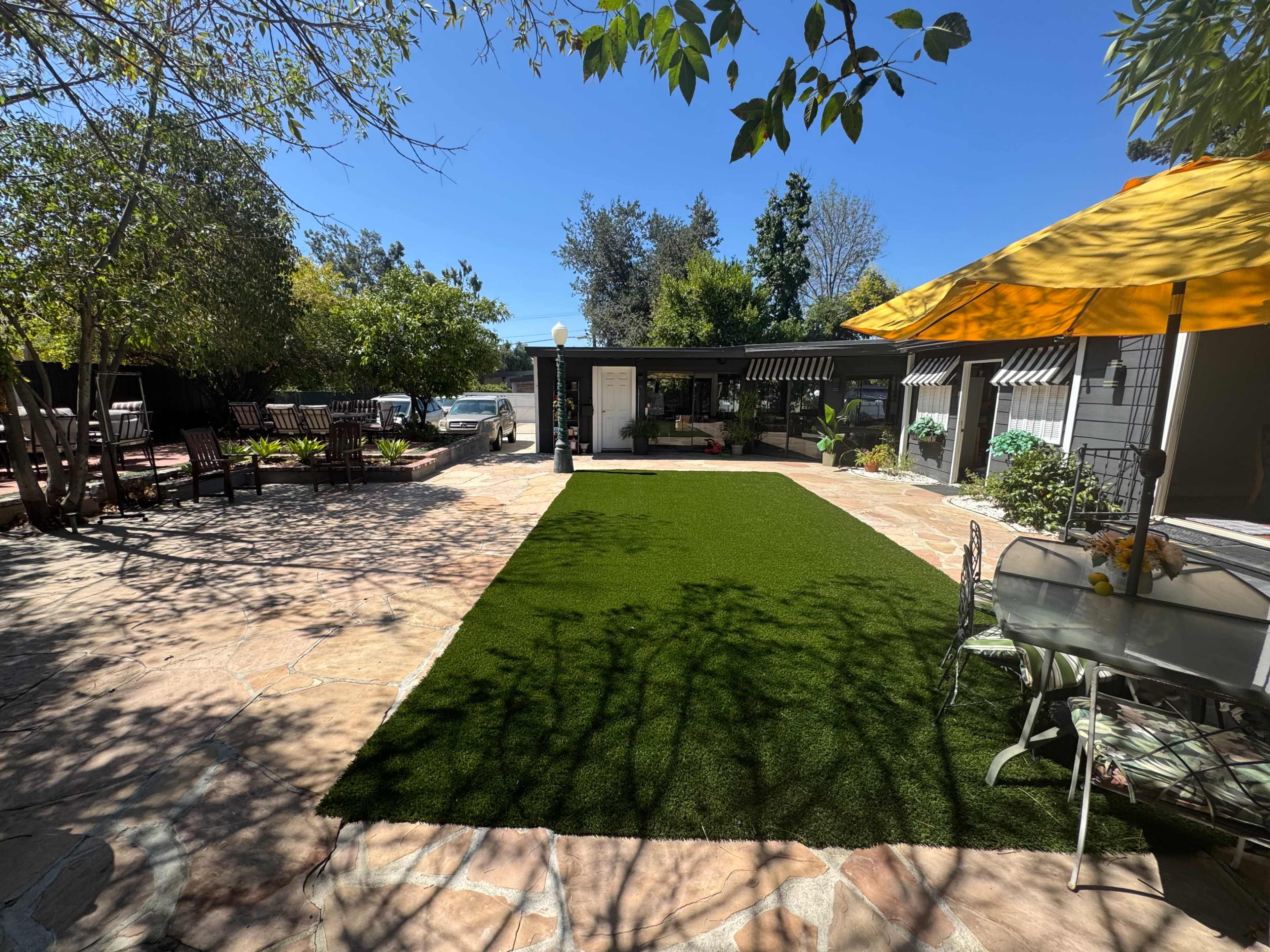 The image shows a landscaped outdoor space featuring a green artificial lawn surrounded by stone pathways, outdoor furniture, and trees under a clear blue sky.