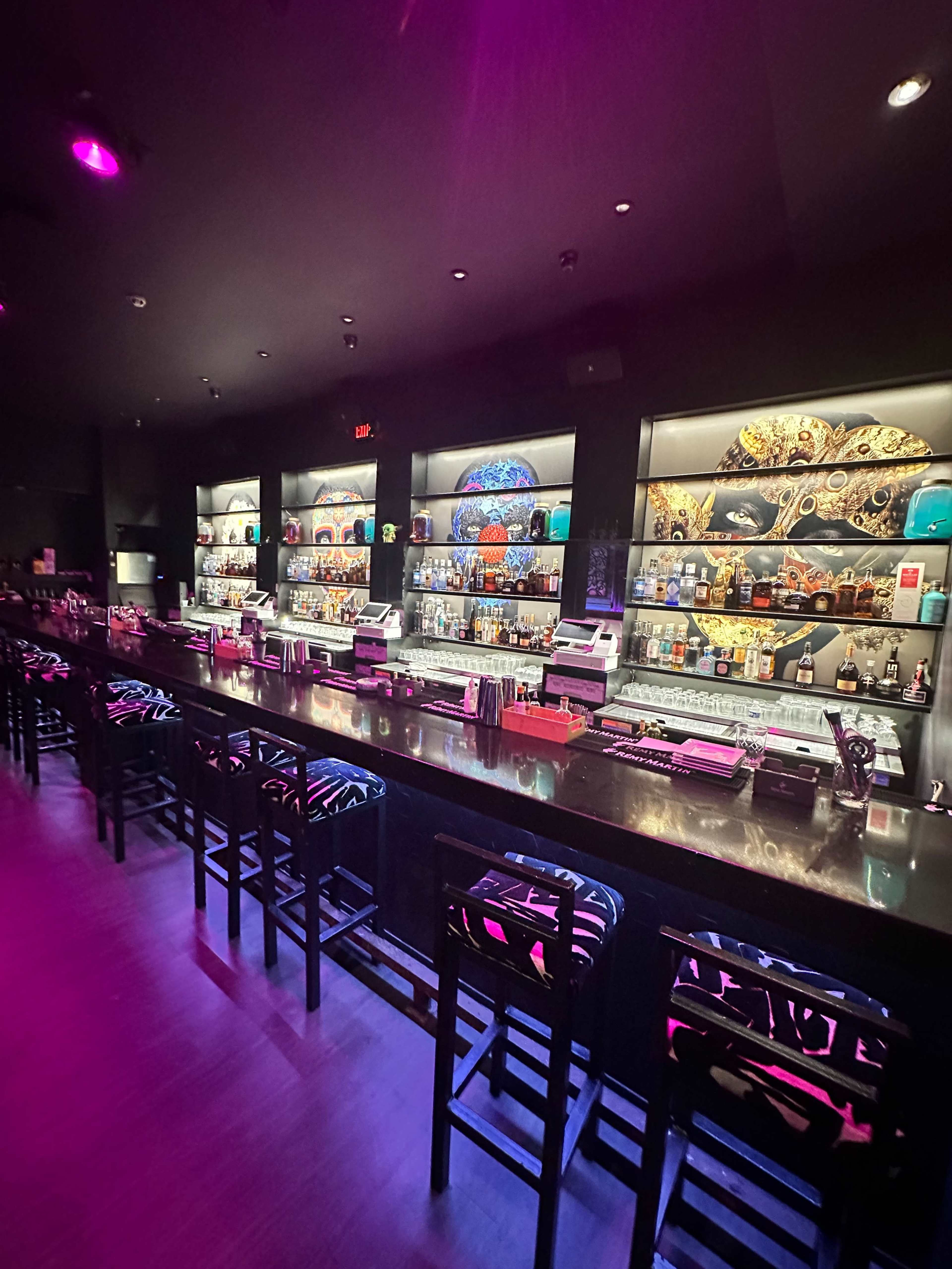 A dimly lit bar with a long countertop, shelves filled with various bottles of liquor, and colorful decorative elements in the background.