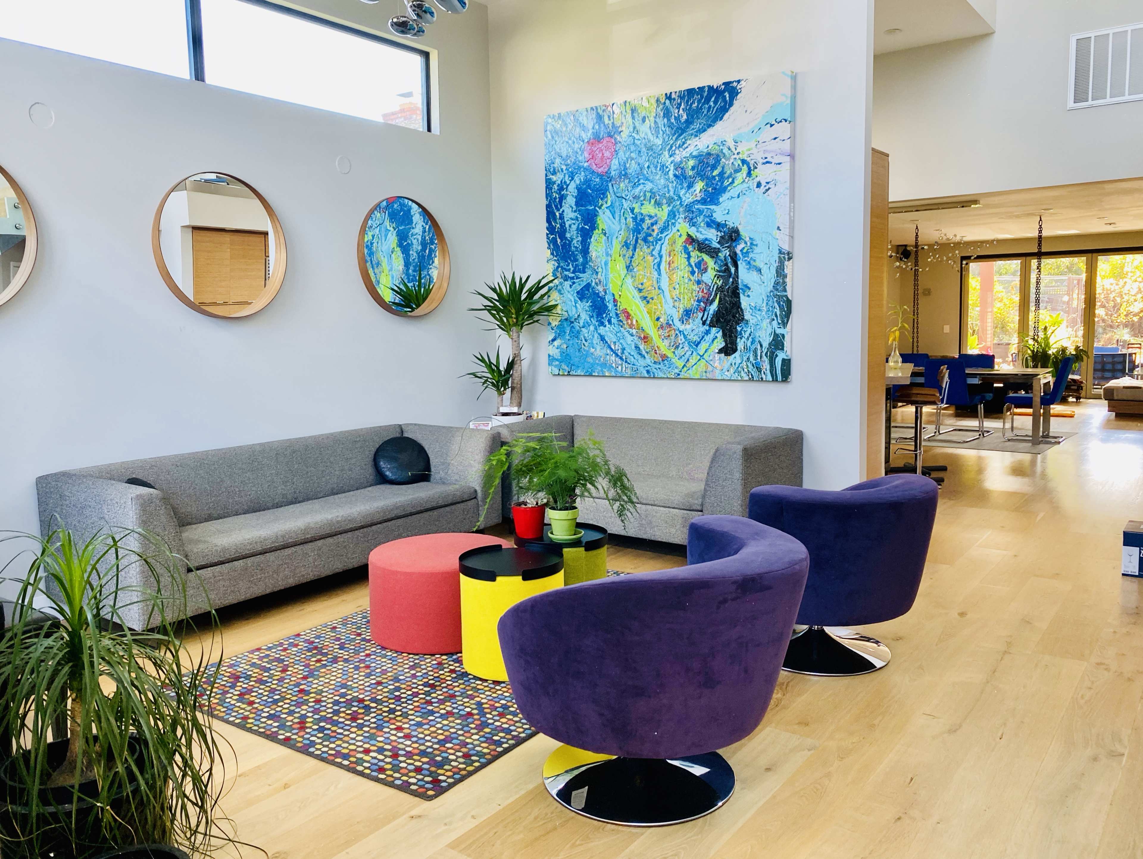 A modern living room with a gray sofa, colorful round stools, a large abstract painting, and potted plants, all set in a bright, open space.