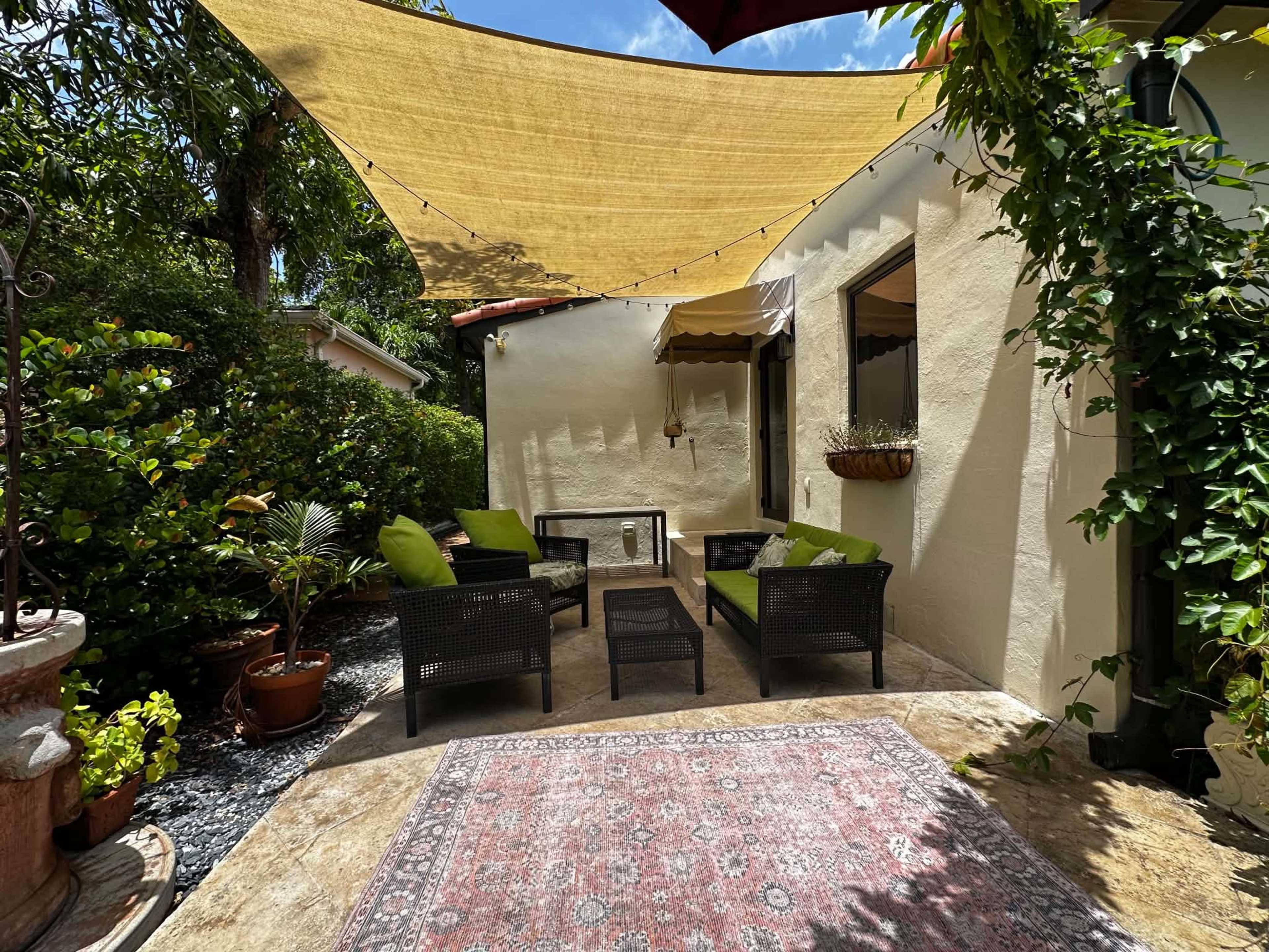 A shaded outdoor seating area features black wicker chairs with green cushions, a decorative rug, and surrounding plants.