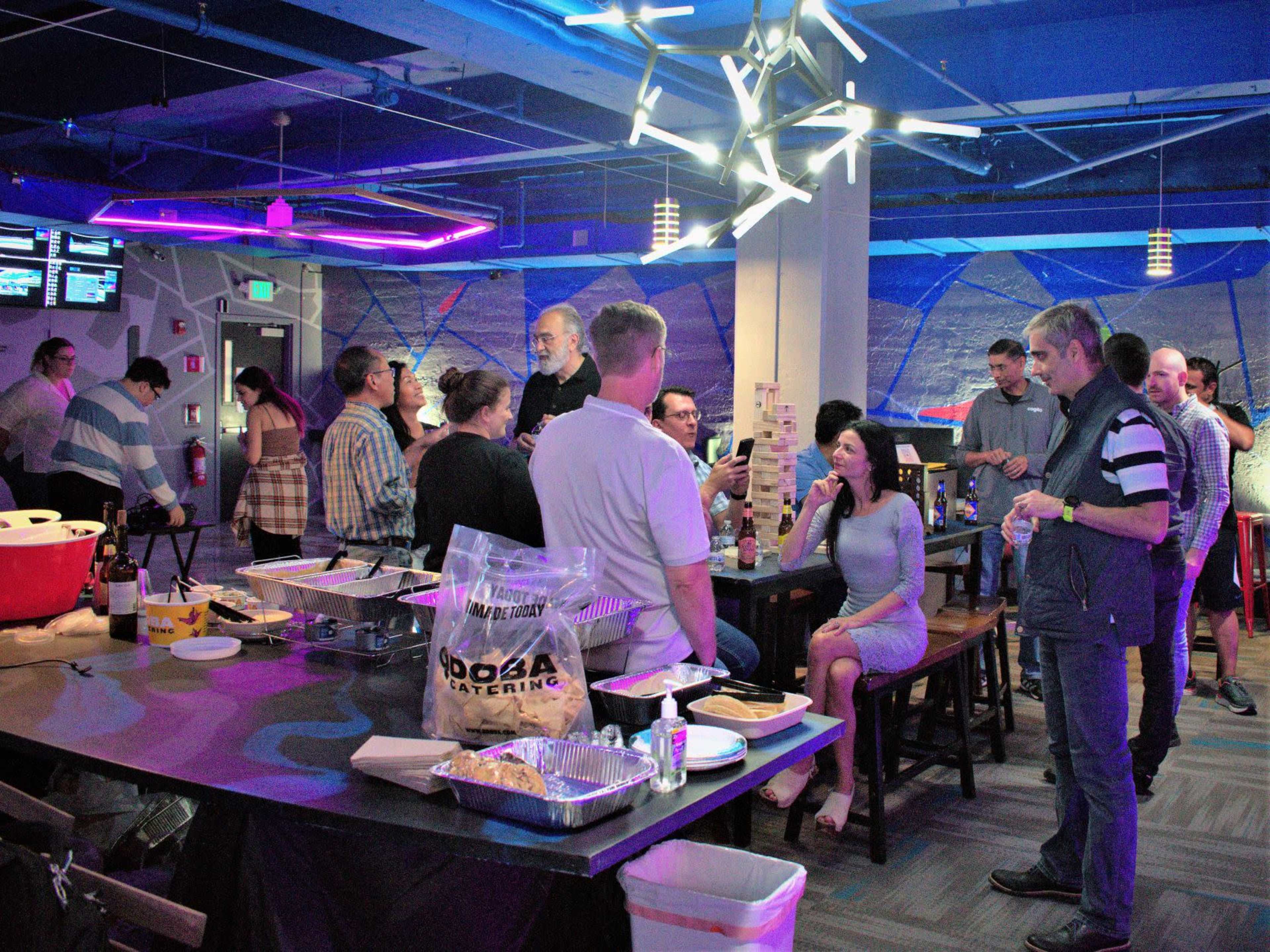 A group of people socializes in a modern space decorated with vibrant lighting while enjoying food and beverages at a gathering.