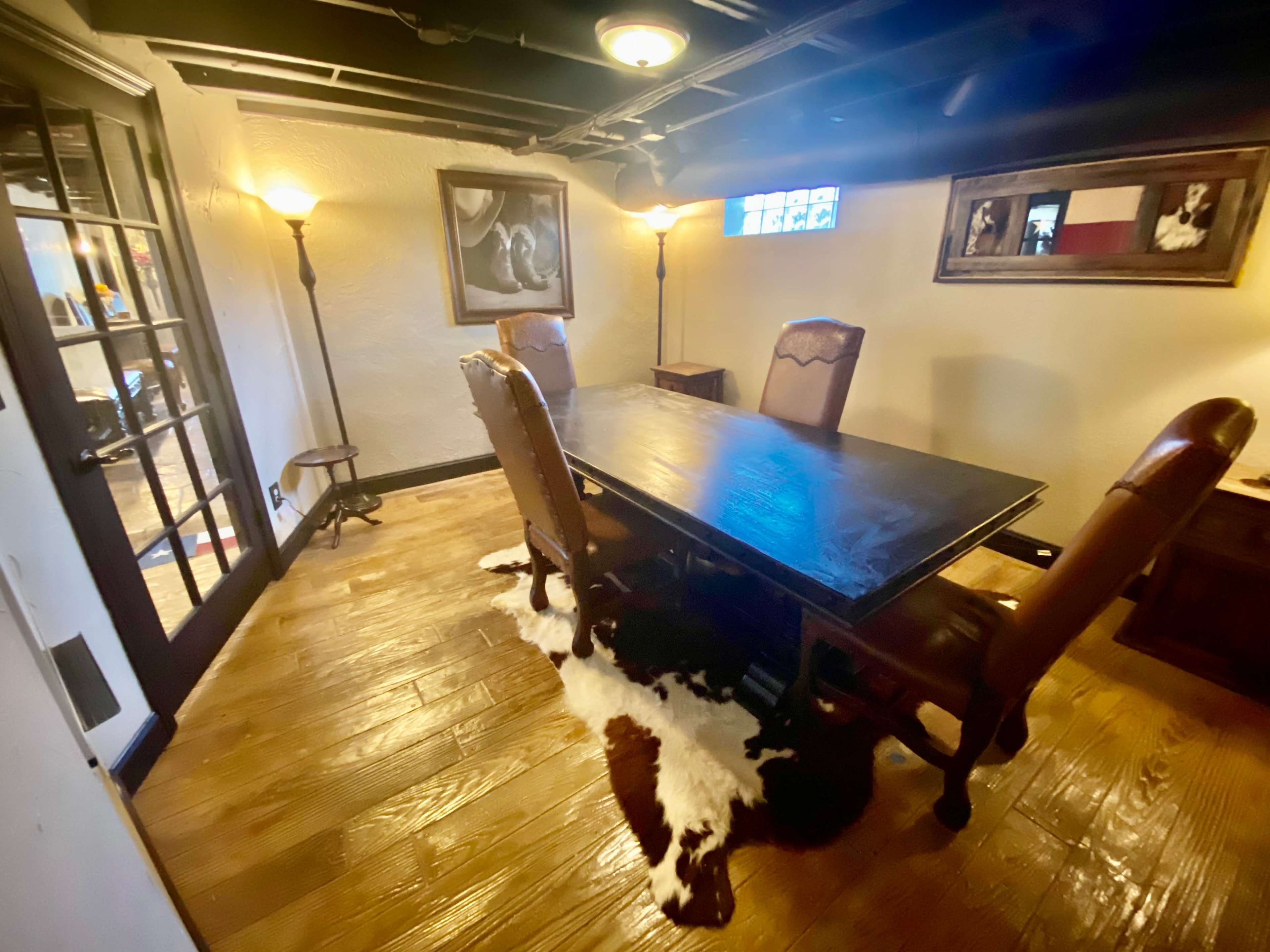 A dimly lit room with a large wooden table surrounded by four leather chairs, a cowhide rug on the floor, and framed artwork on the walls.