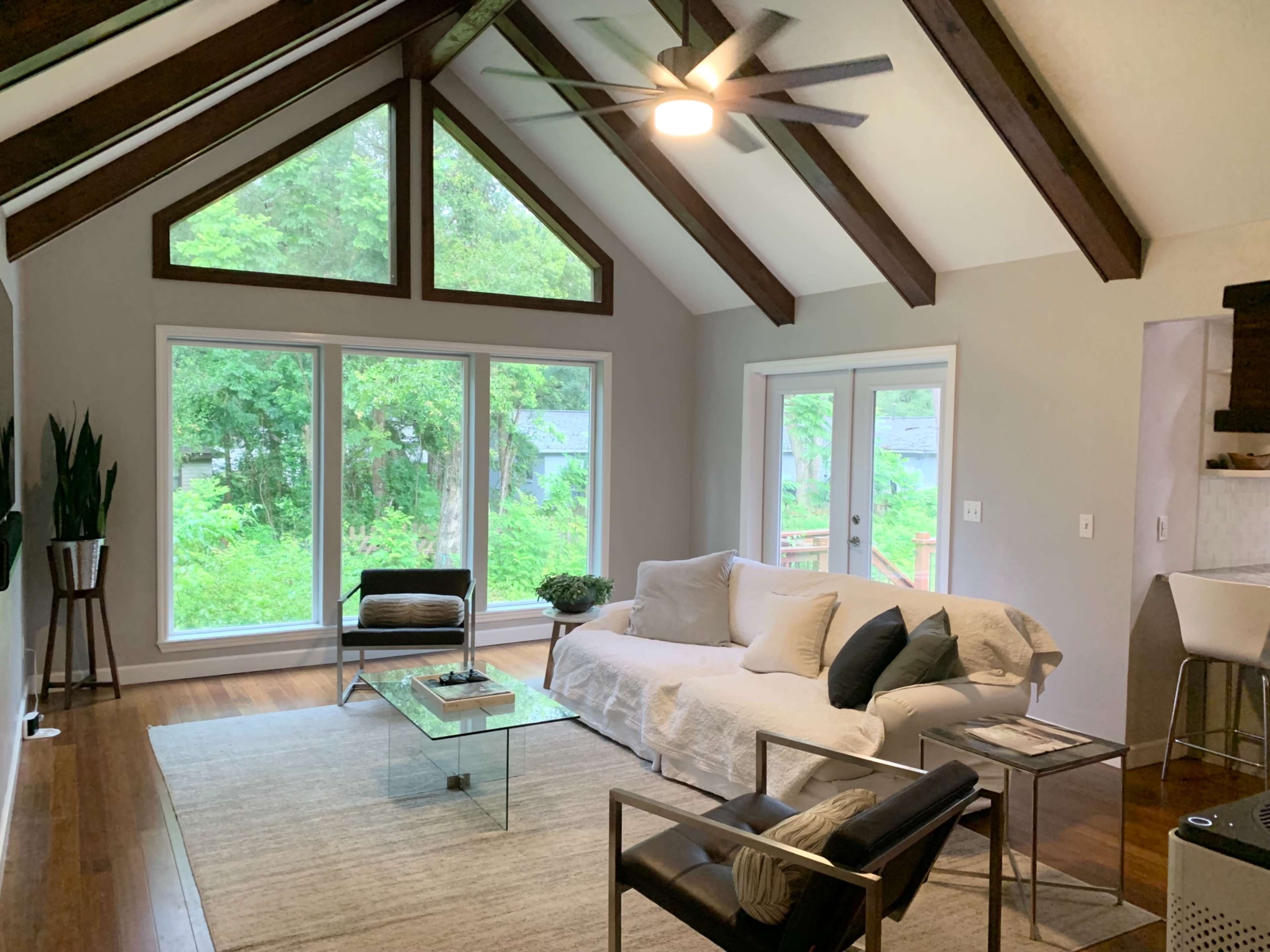 The image shows a modern living room featuring a large window with a view of greenery, a light-colored sofa, a glass coffee table, and wooden beams on the ceiling.