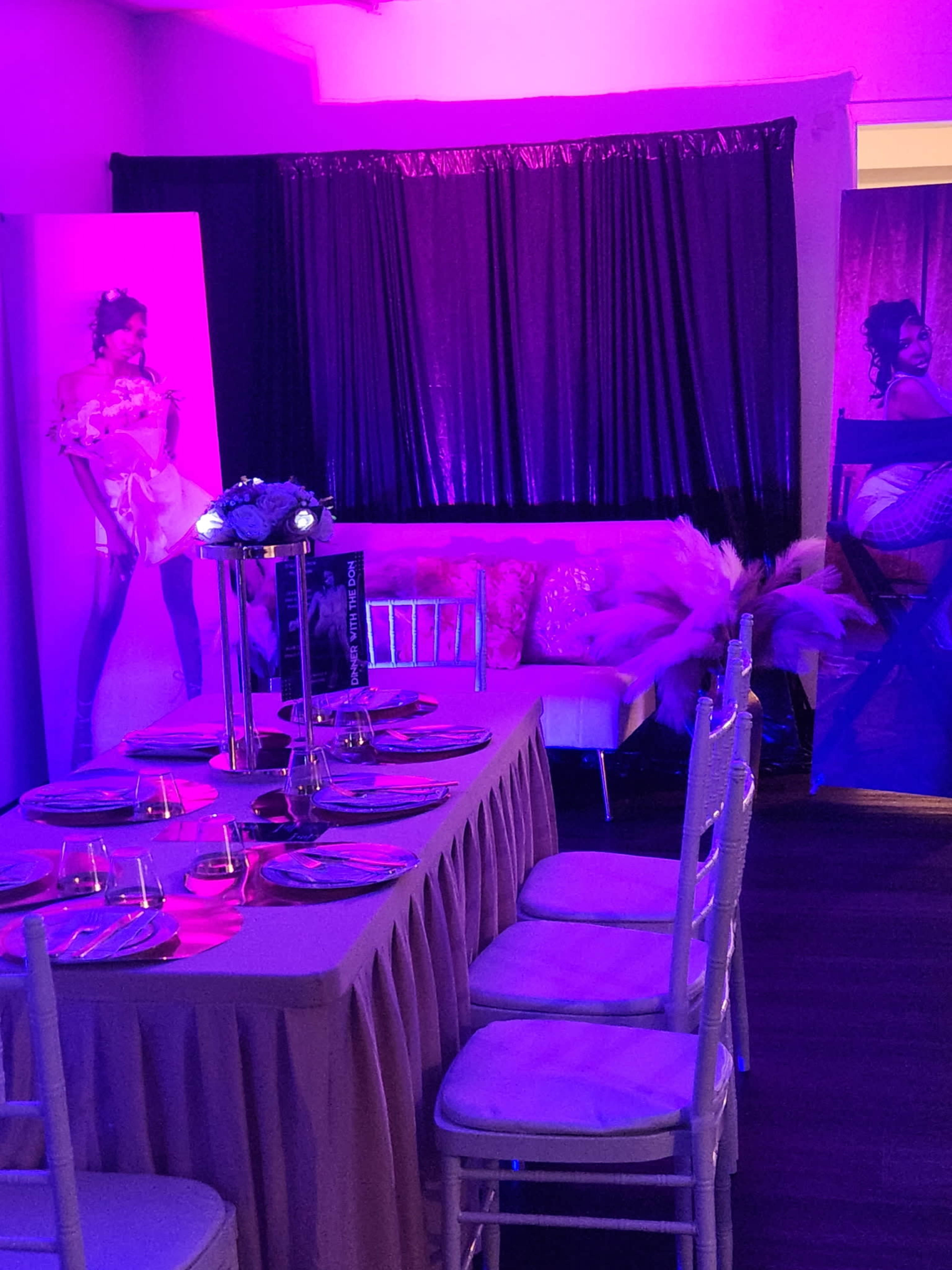 The image shows an elegantly decorated party space with a long table set for dining, adorned with plates, and flanked by tall candle holders, surrounded by vibrant lighting and decorative elements.
