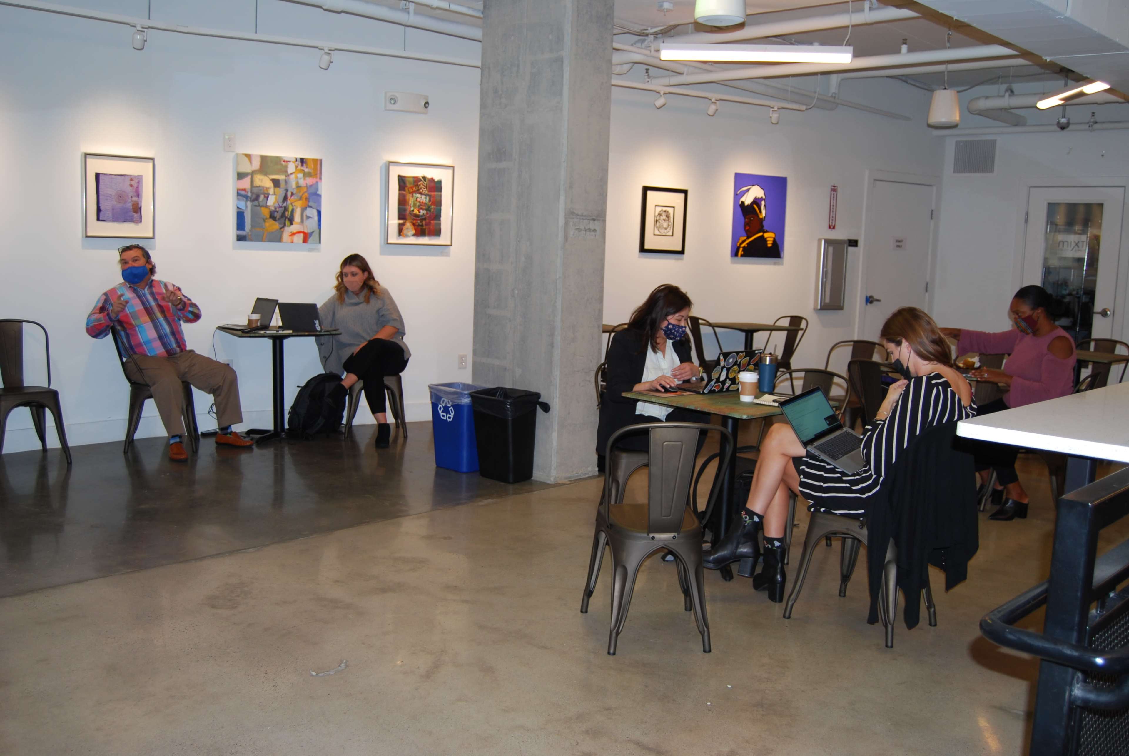 The image shows a spacious room with several people working at tables, wearing masks, and various artworks displayed on the walls.