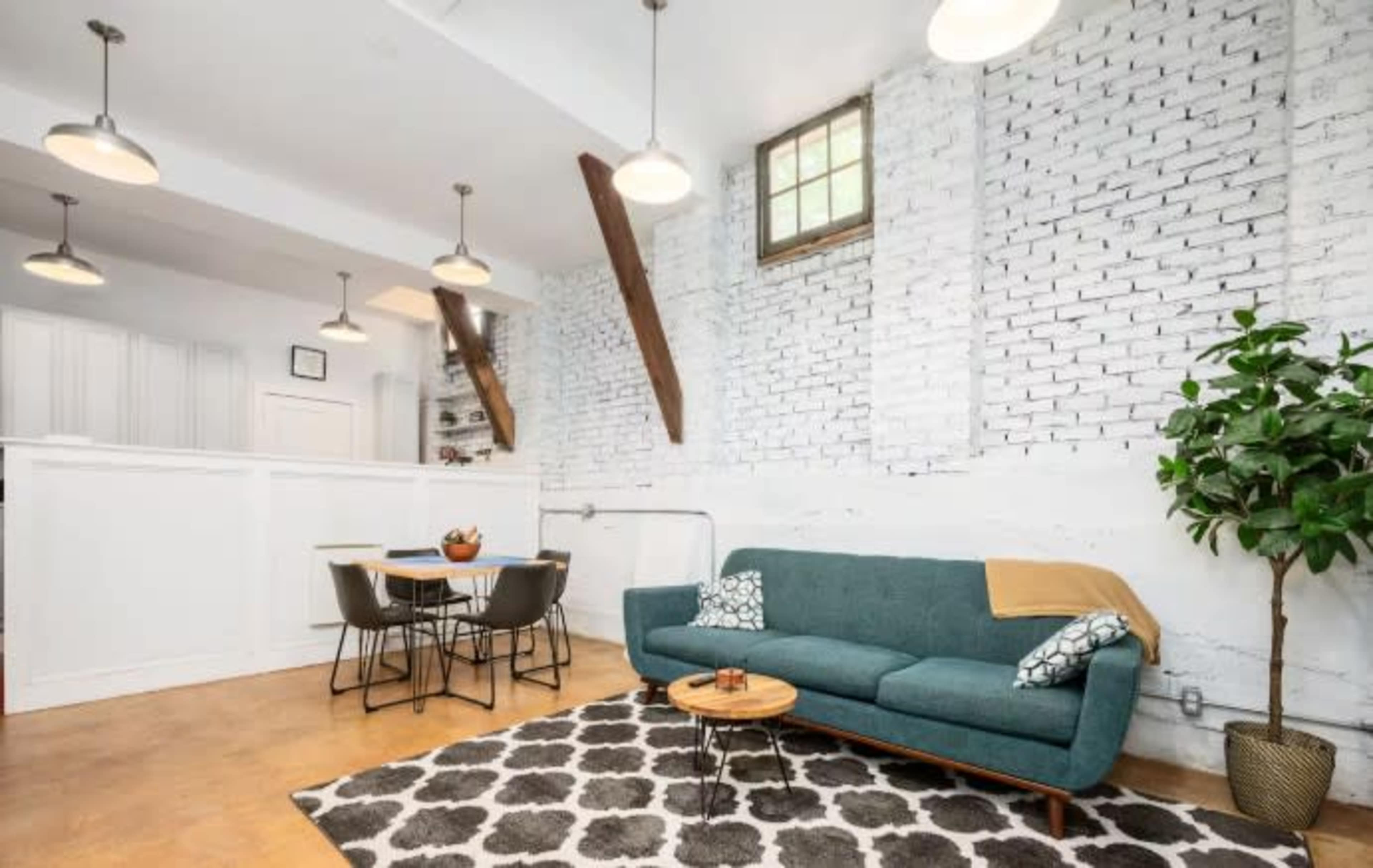 The image shows a modern living space featuring a teal sofa, a round wooden coffee table, and a dining area with black chairs, all set against a backdrop of white brick walls and wooden beams.