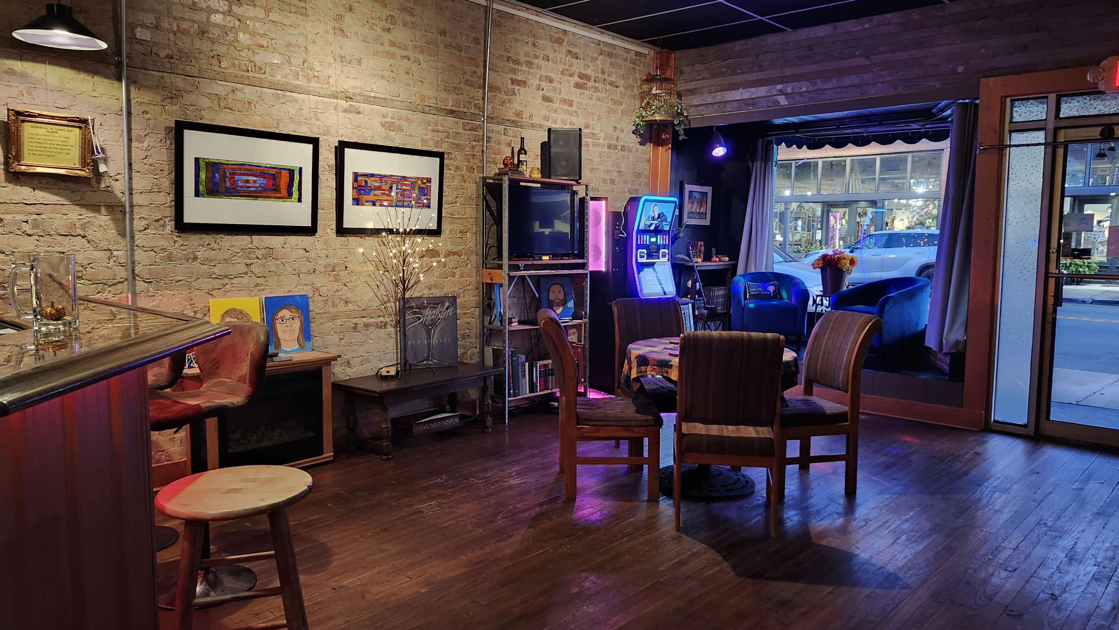 The interior of a cozy bar features exposed brick walls, wooden flooring, and a mix of seating arrangements, with colorful artwork displayed on the walls.