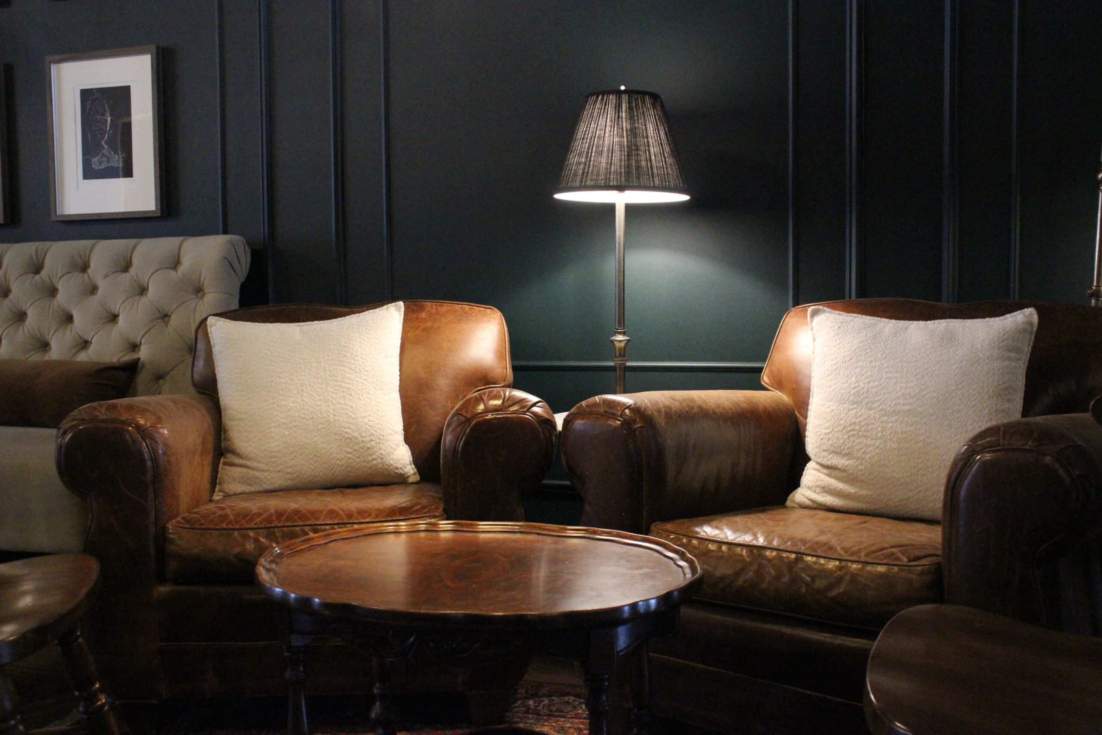 The scene features two leather armchairs with decorative pillows flanking a round wooden coffee table, illuminated by a lamp in a dark-walled room.