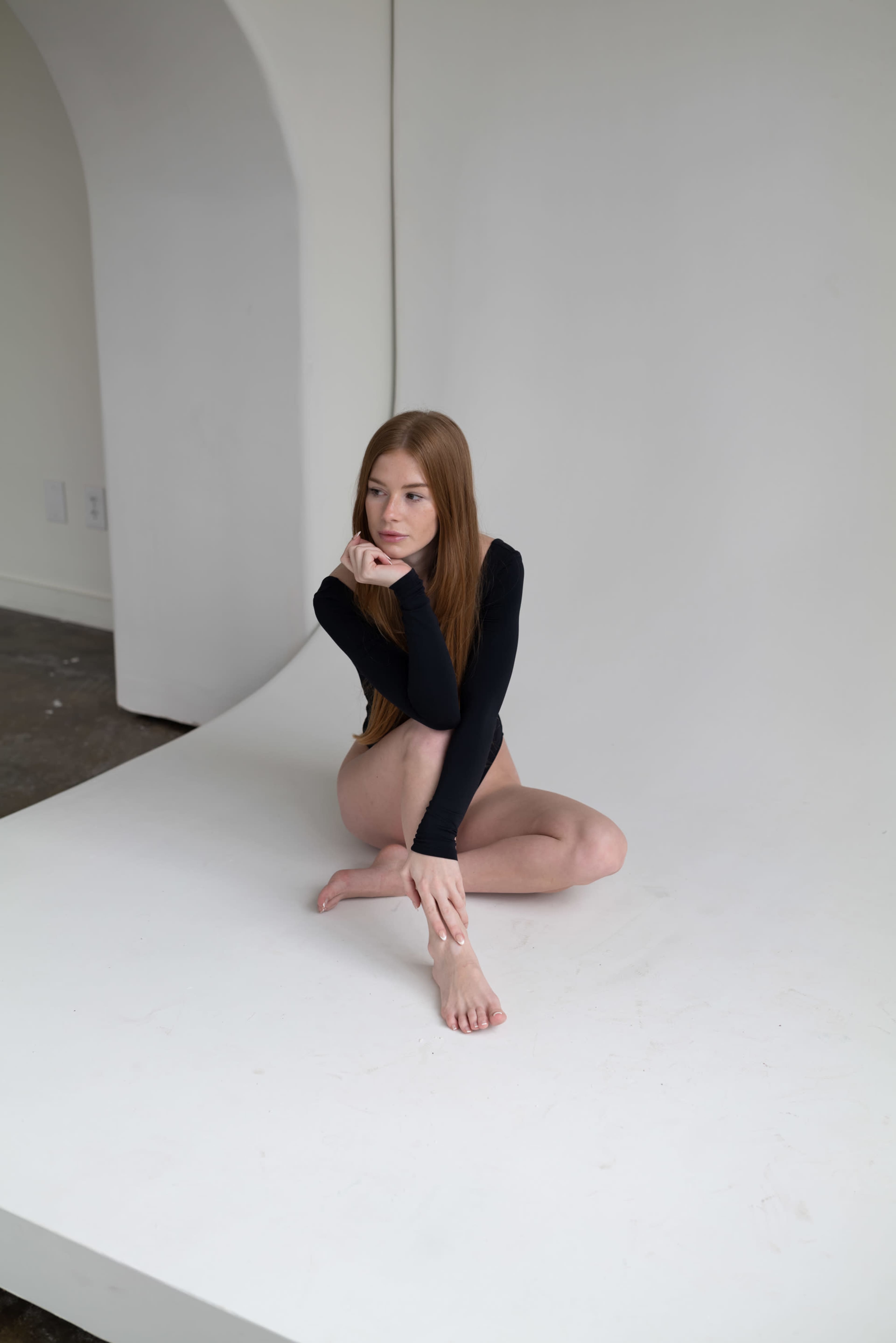 A person with long hair is sitting on a white backdrop, resting their chin on one hand with their knees drawn up to their chest.