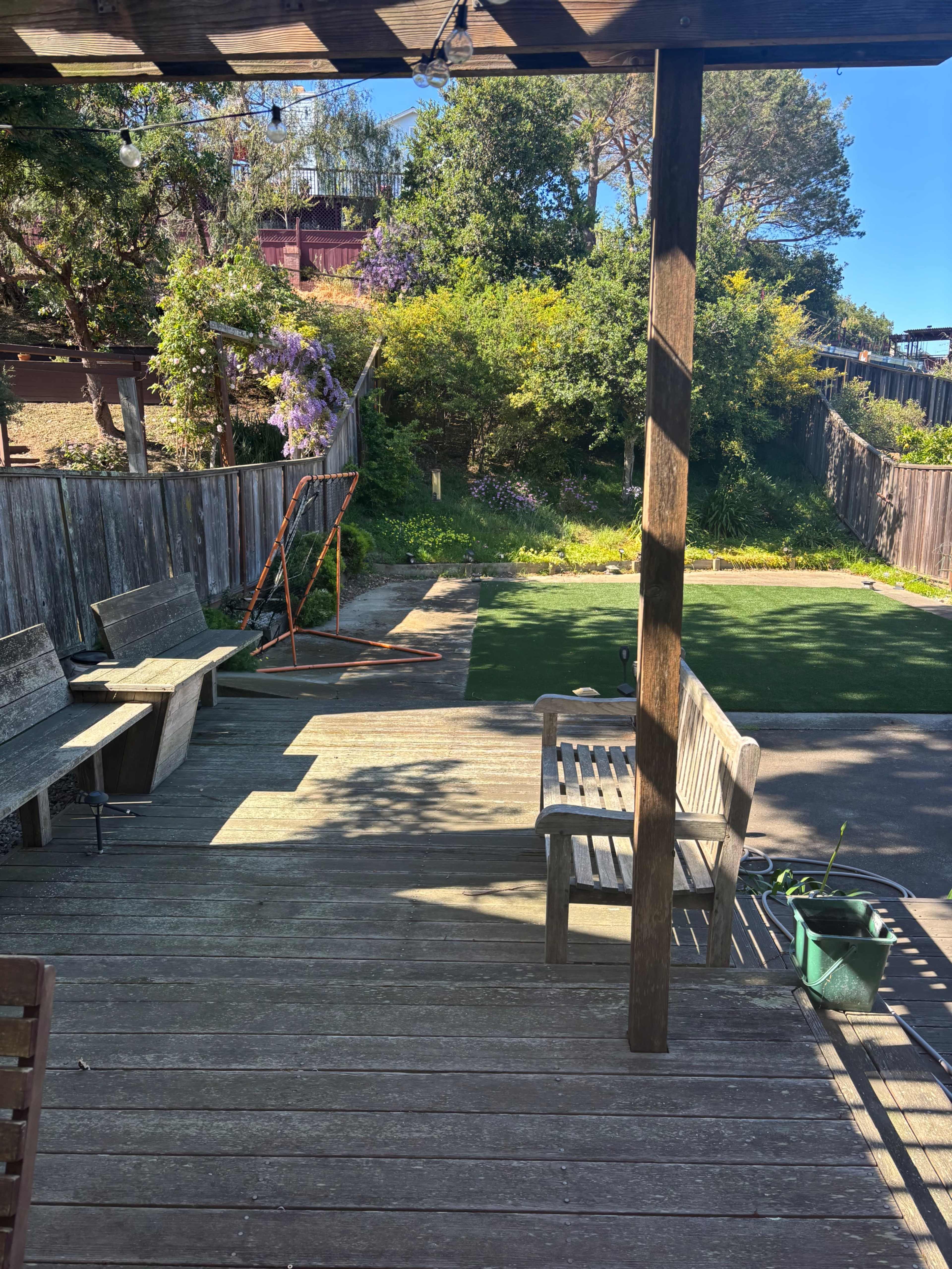 The image shows a wooden deck with benches, a green lawn area, and a swing set in a backyard surrounded by trees and a wooden fence.