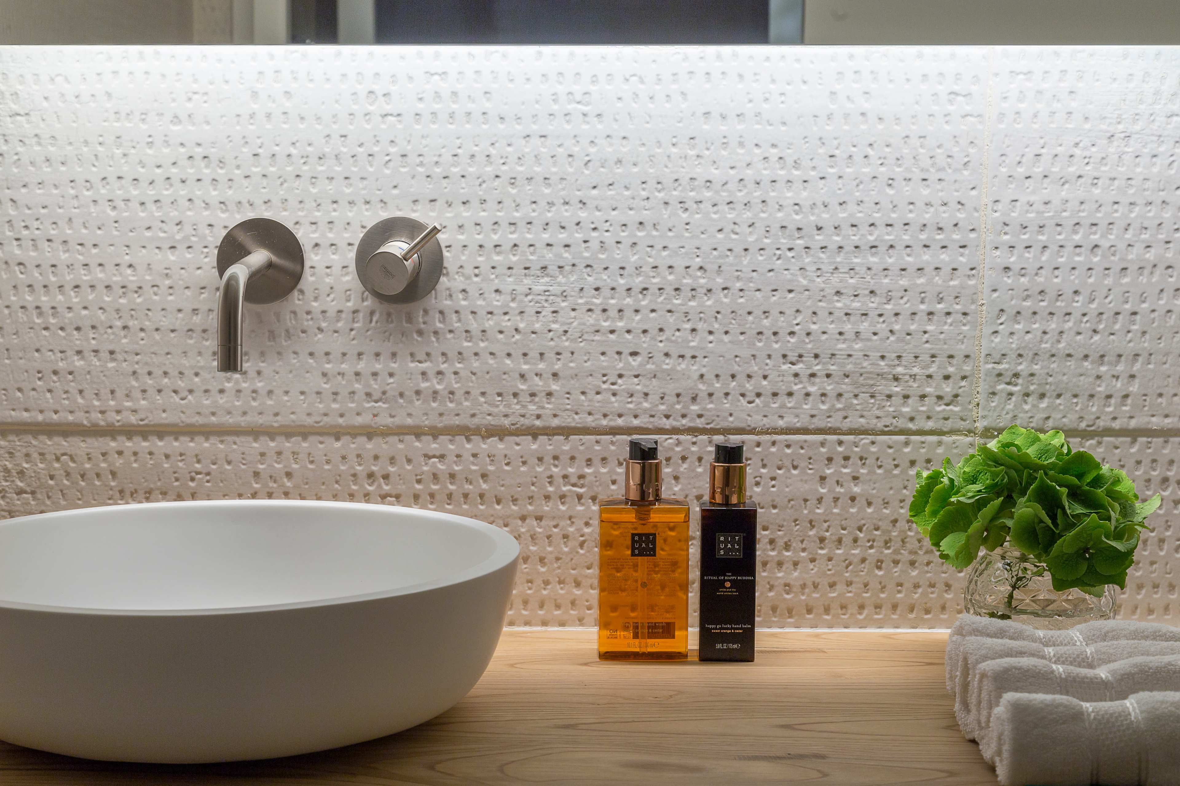 A modern bathroom with a round white sink, two bottles of liquid soap in amber and black, and a small green plant beside a folded towel on a wooden surface.
