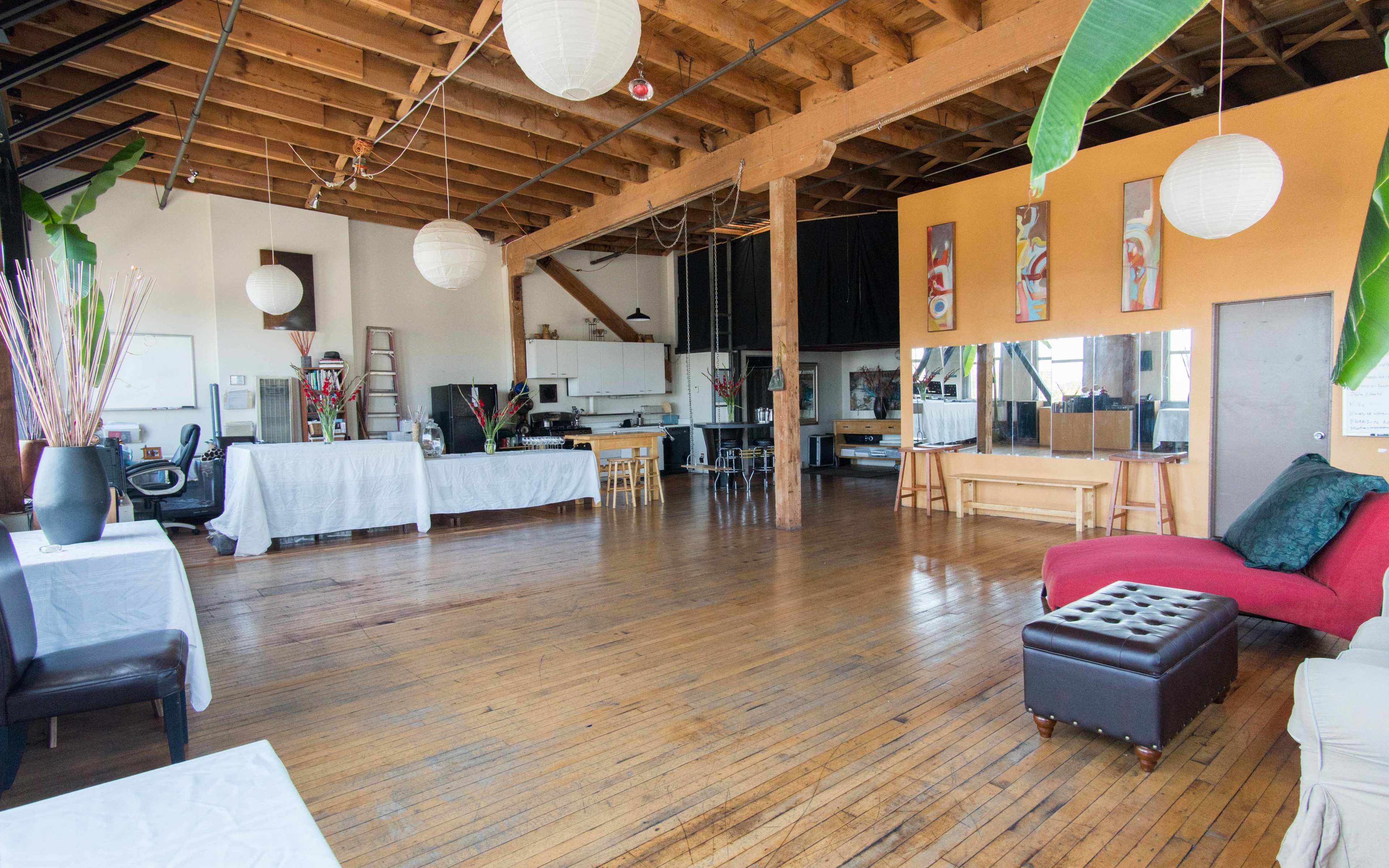 A spacious open room with wooden flooring, high ceilings, and a kitchen area in the background, furnished with tables, chairs, and decorative elements.