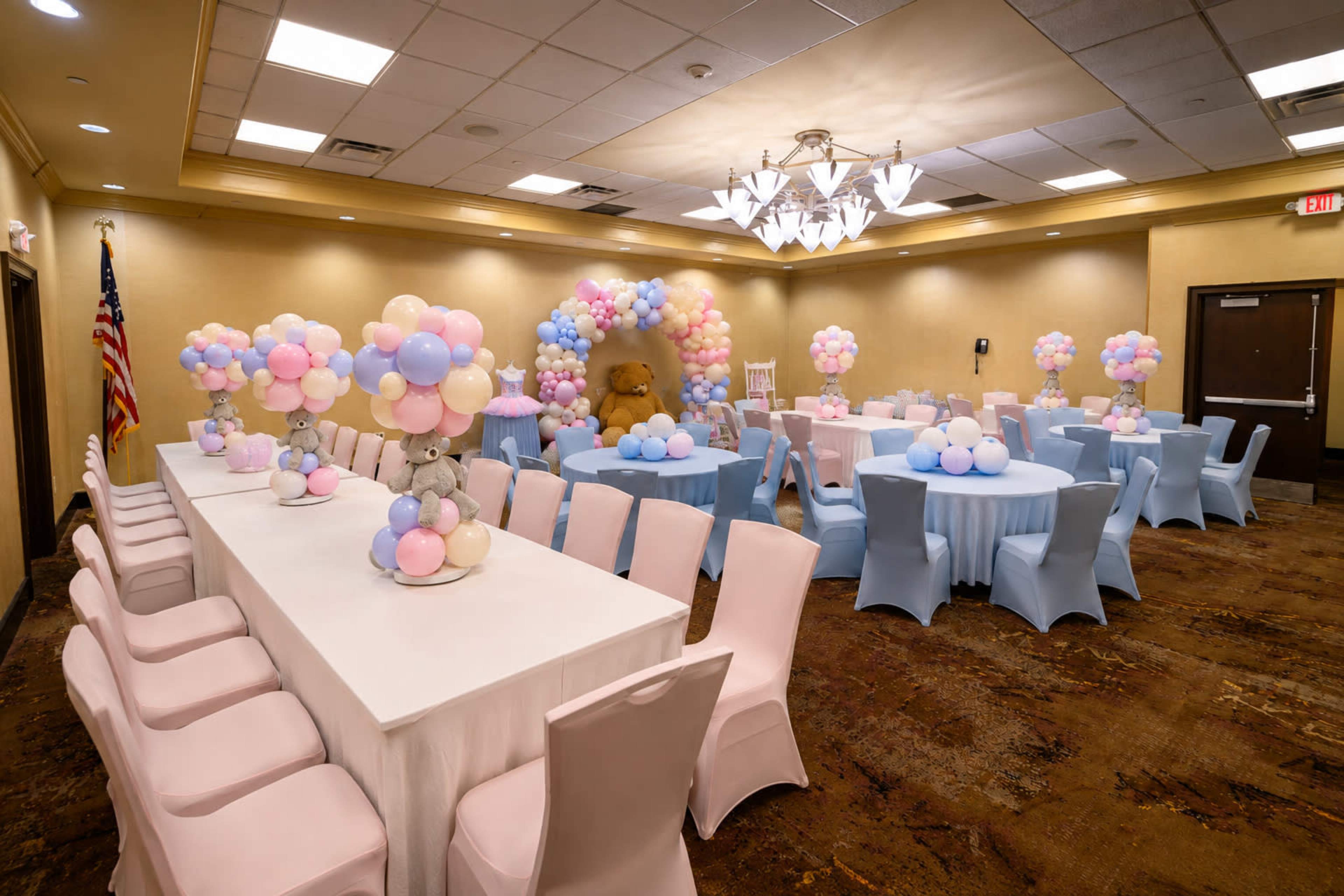 The image shows a decorated event space featuring tables covered in white and light blue tablecloths and centerpieces of pastel balloons, with an elaborate balloon arch in the background.