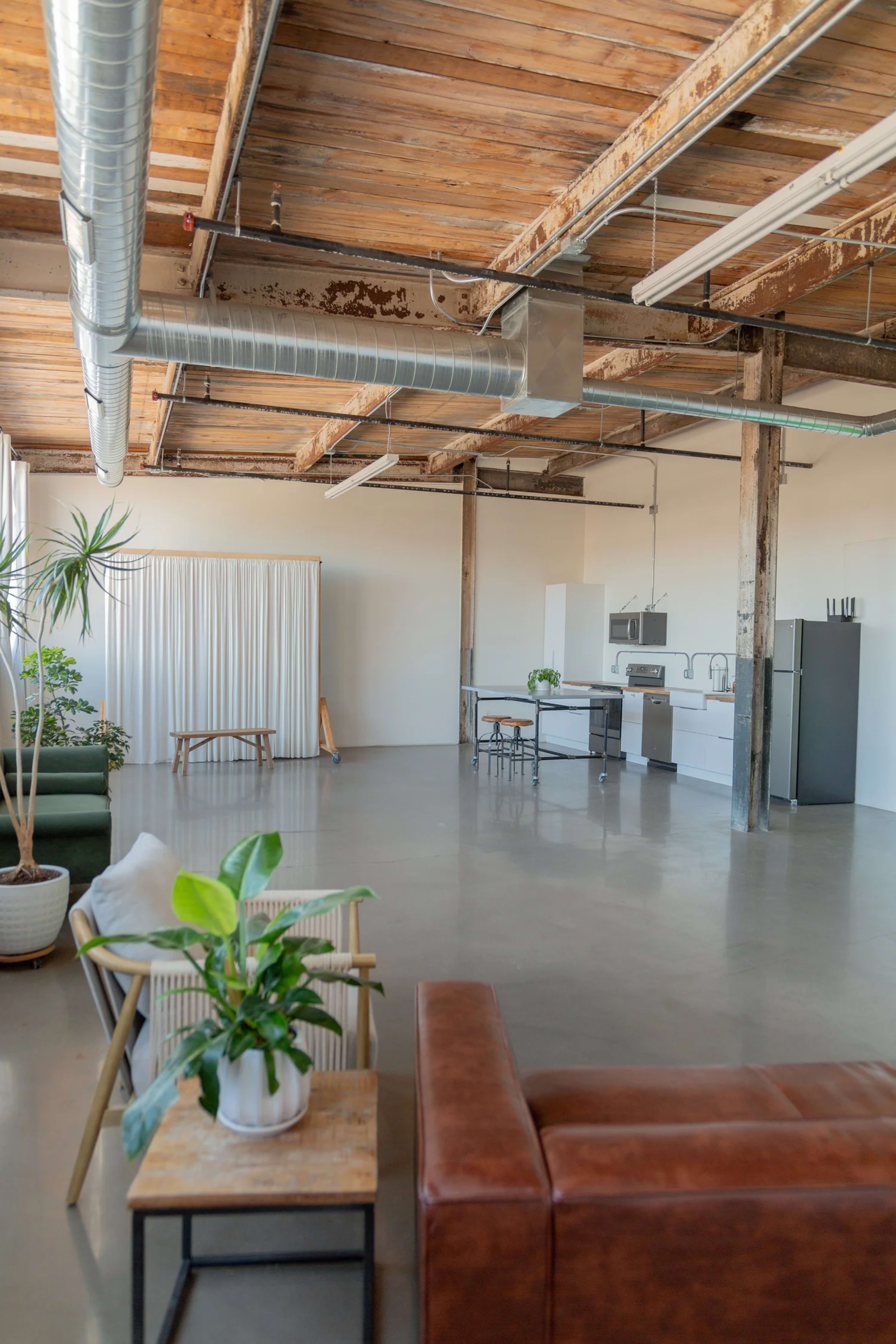 The image shows a spacious, industrial-style loft with exposed wooden beams, polished concrete floors, and a minimalistic kitchen in the background.
