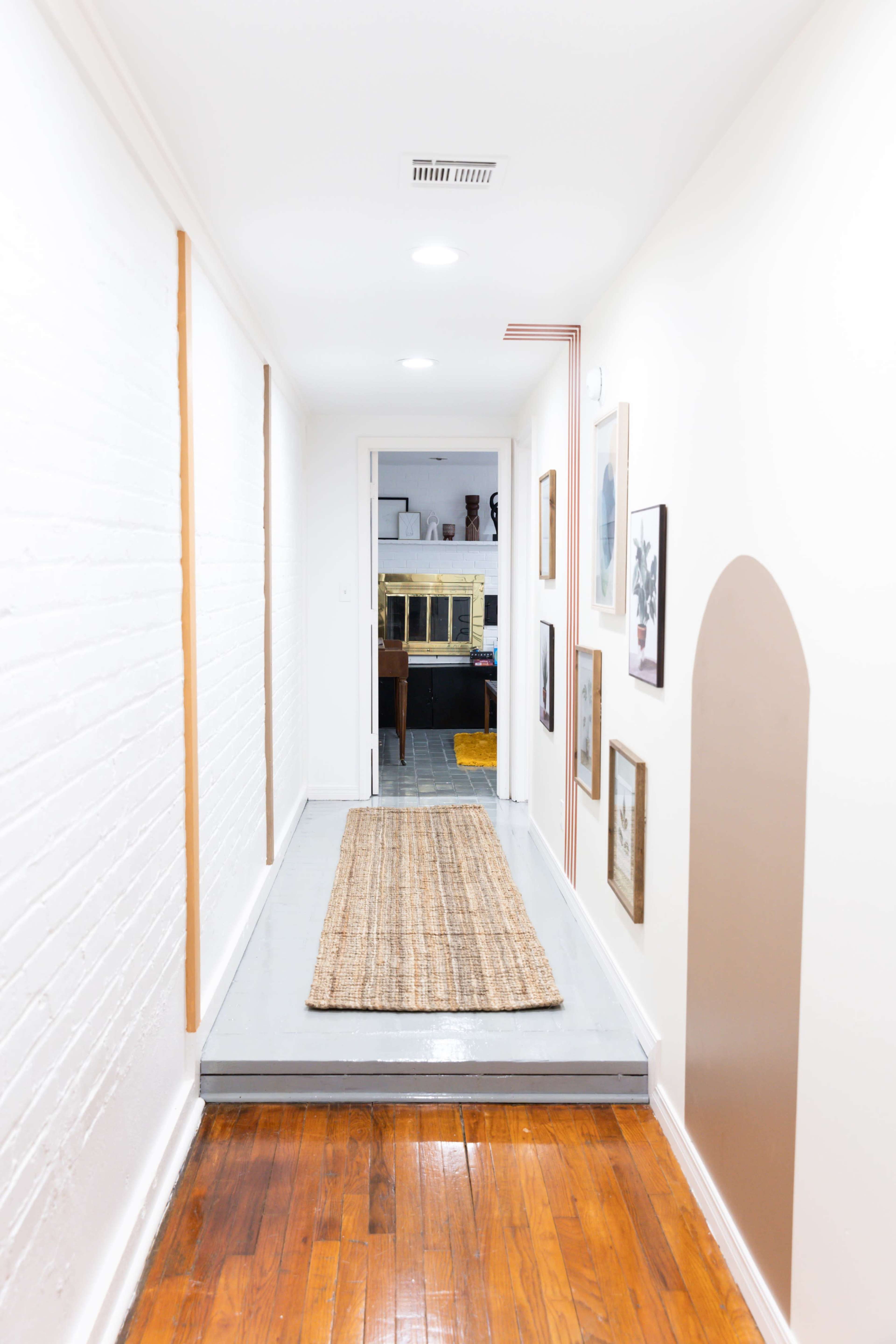 A narrow hallway features a woven rug and framed artwork on the walls, leading to a doorway at the end.