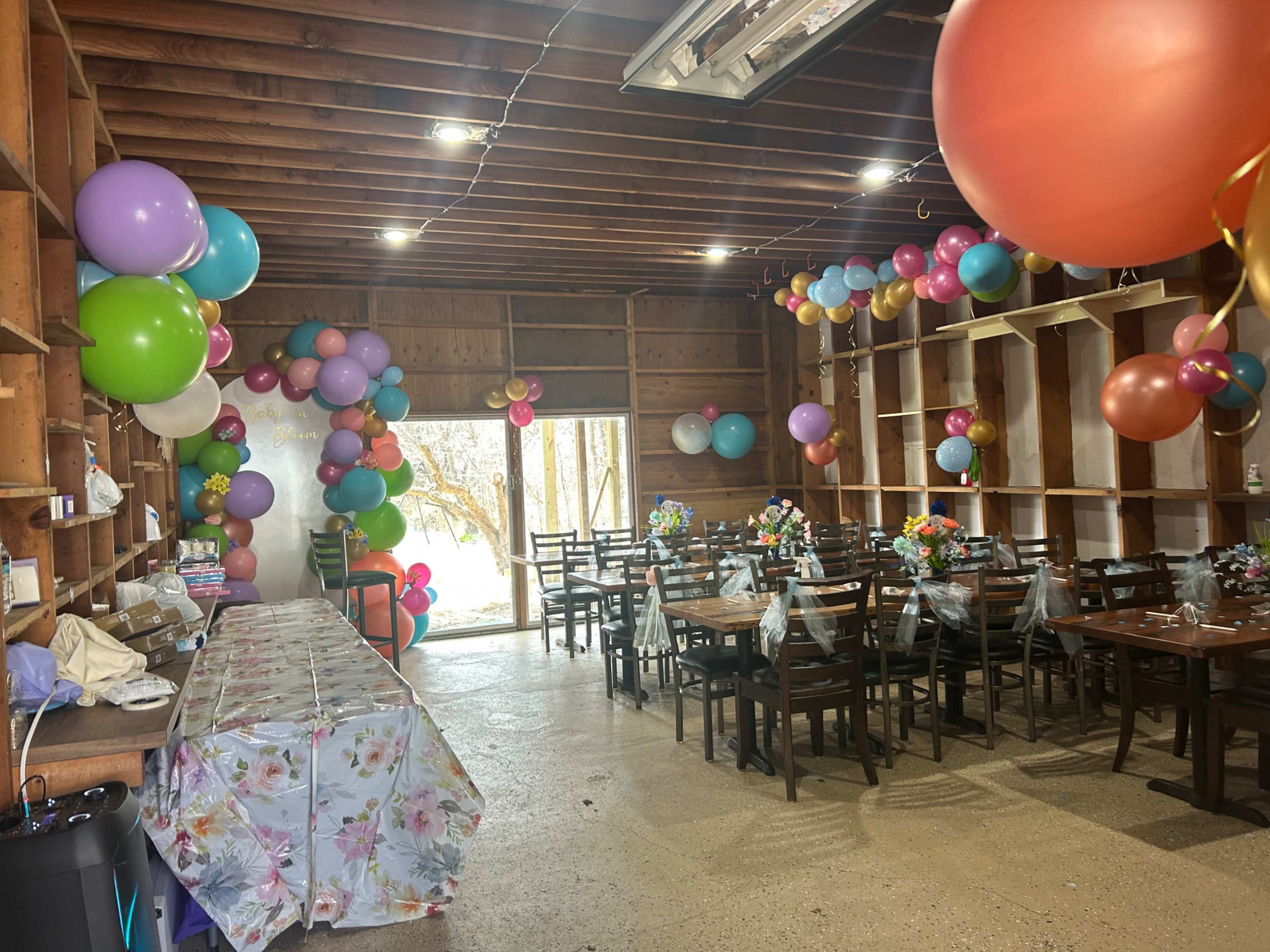 A decorated indoor space with tables and chairs set up, surrounded by colorful balloons and floral arrangements.