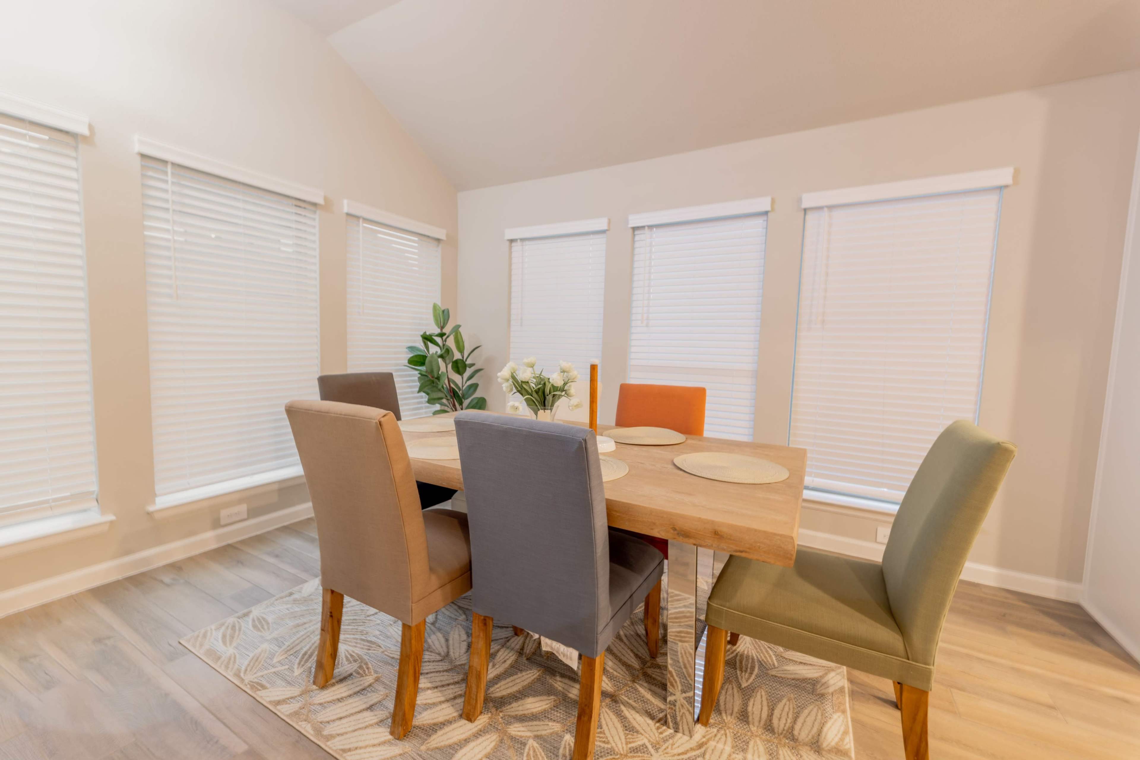 A dining room features a wooden table surrounded by four chairs of different colors, with natural light coming through multiple blinds-covered windows.