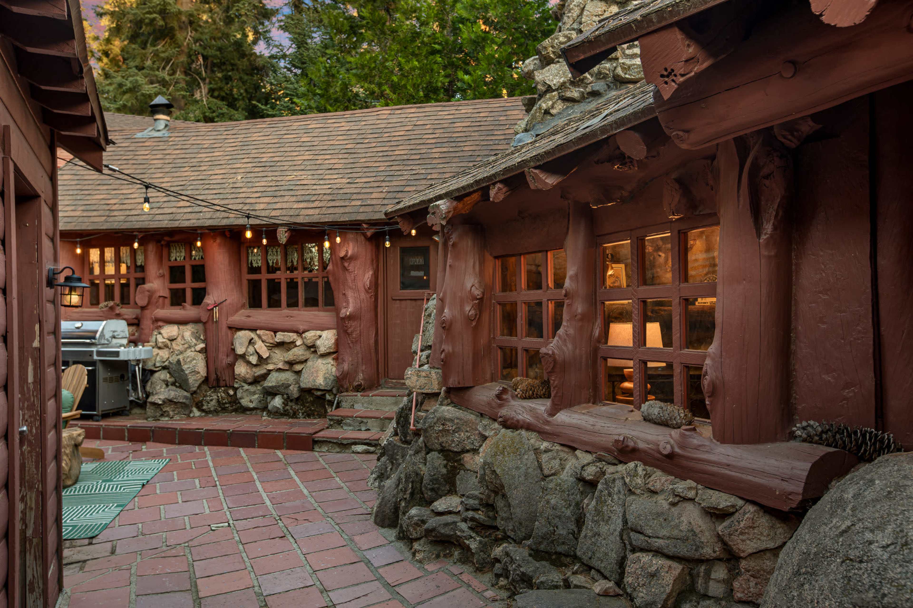 The image shows a rustic courtyard with a stone walkway, surrounded by wooden structures featuring large windows and textured walls.