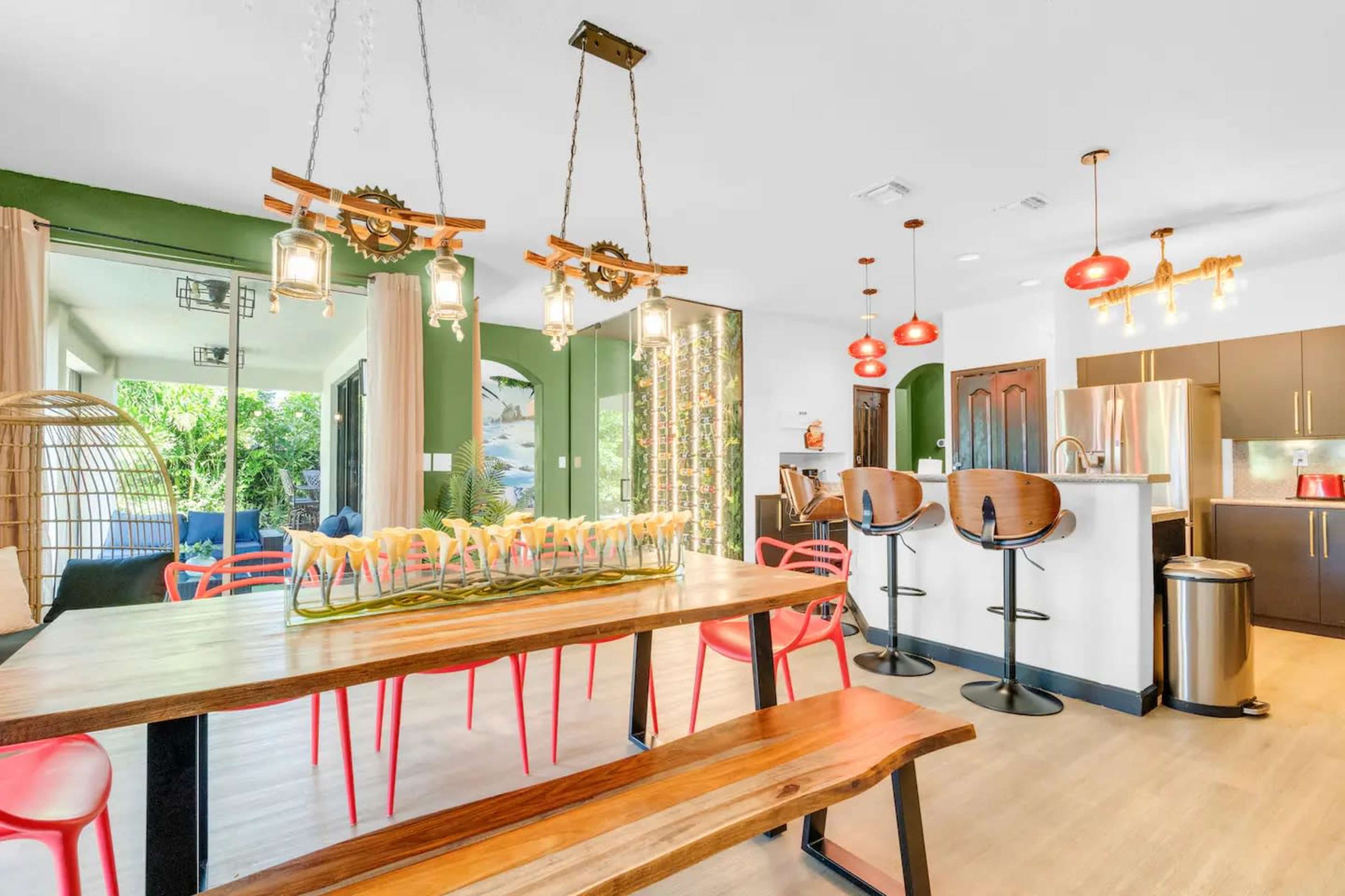 A modern kitchen and dining area features a large wooden table with red chairs, industrial-style lighting, and a bar with stools.