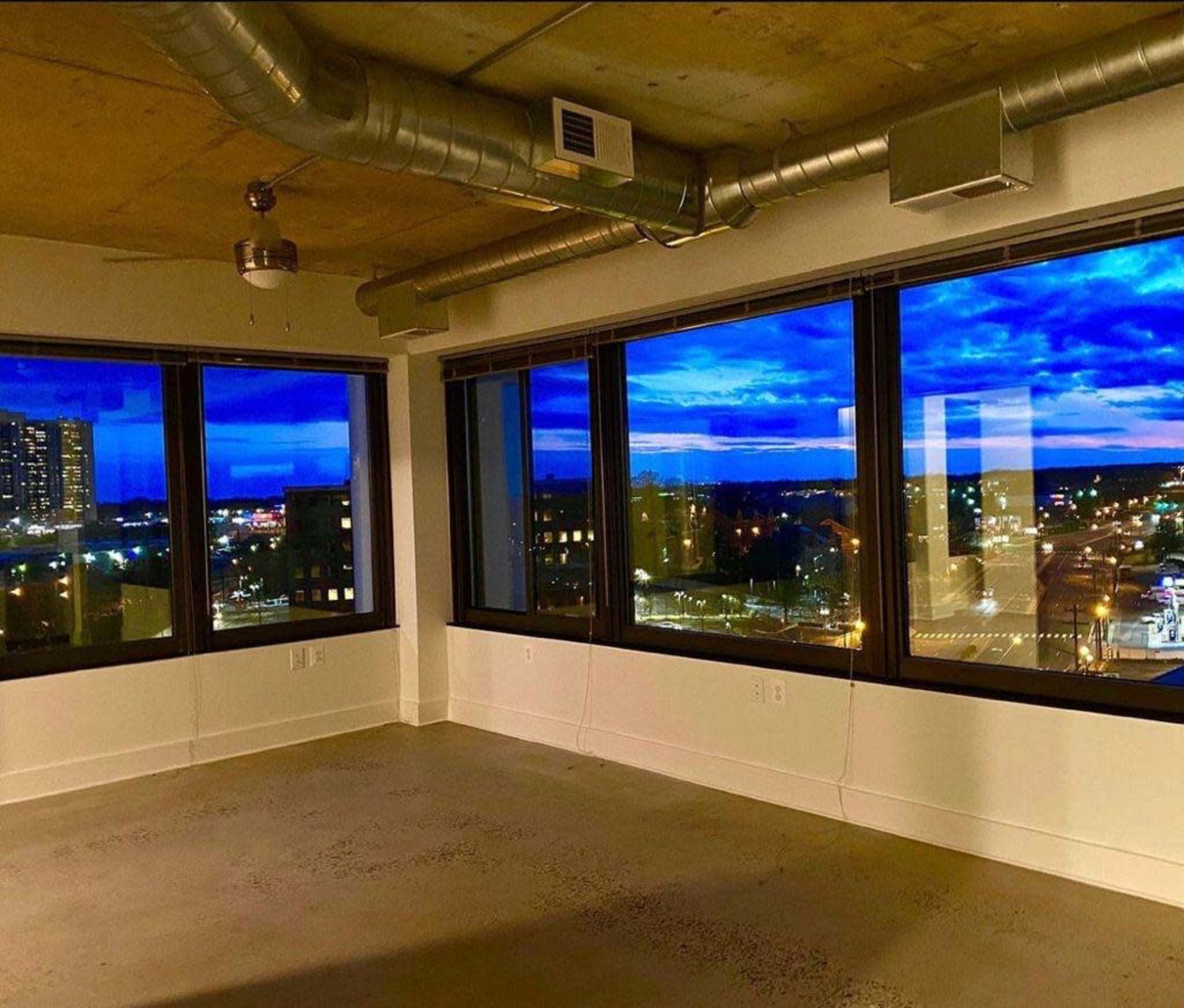 A vacant room with large windows overlooking a cityscape illuminated by evening lights, against a backdrop of a blue sky filled with clouds.