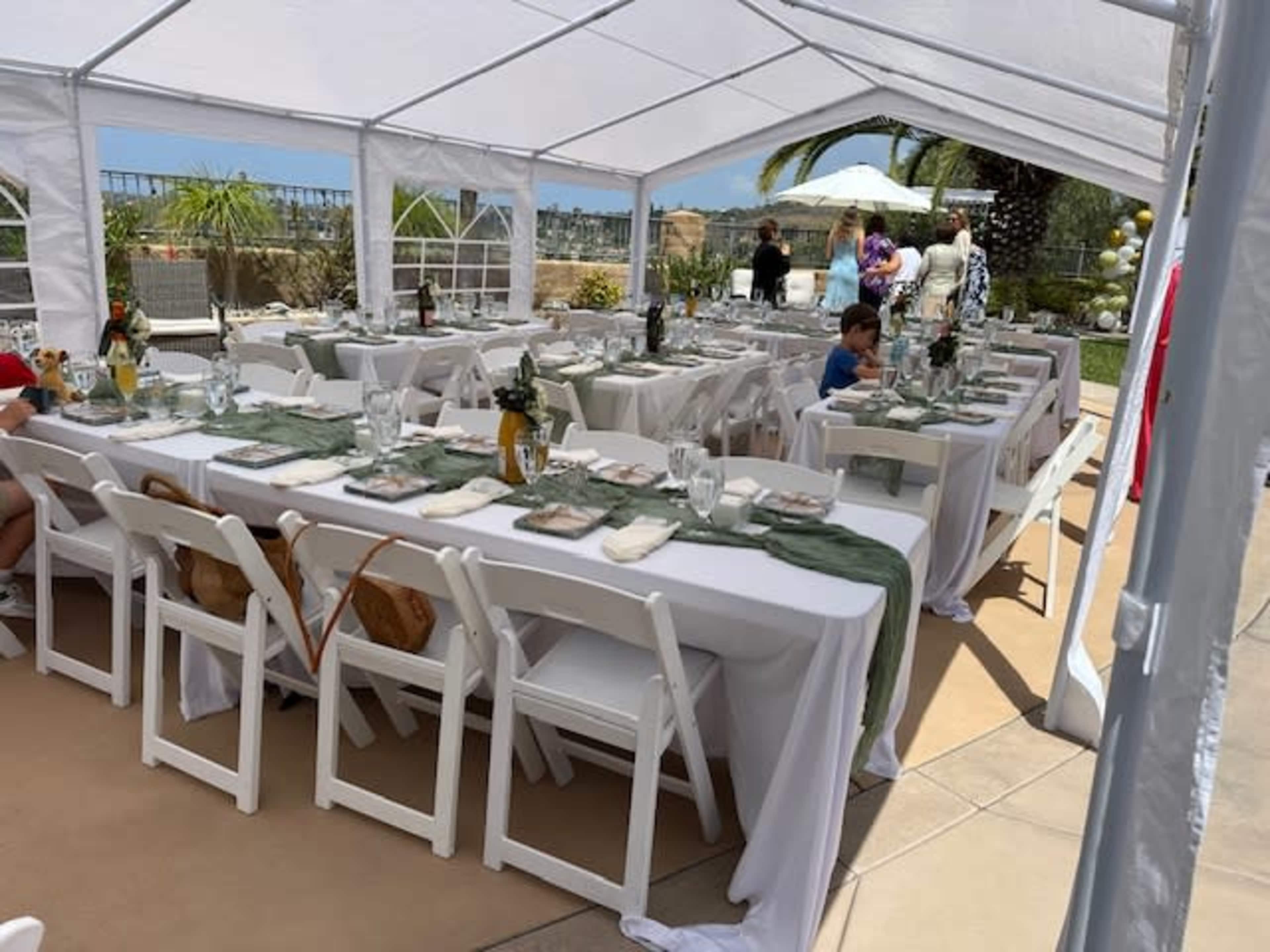 A large outdoor tent is set up with white tables and chairs, decorated with green table runners and glassware, ready for an event, while guests mingle in the background.
