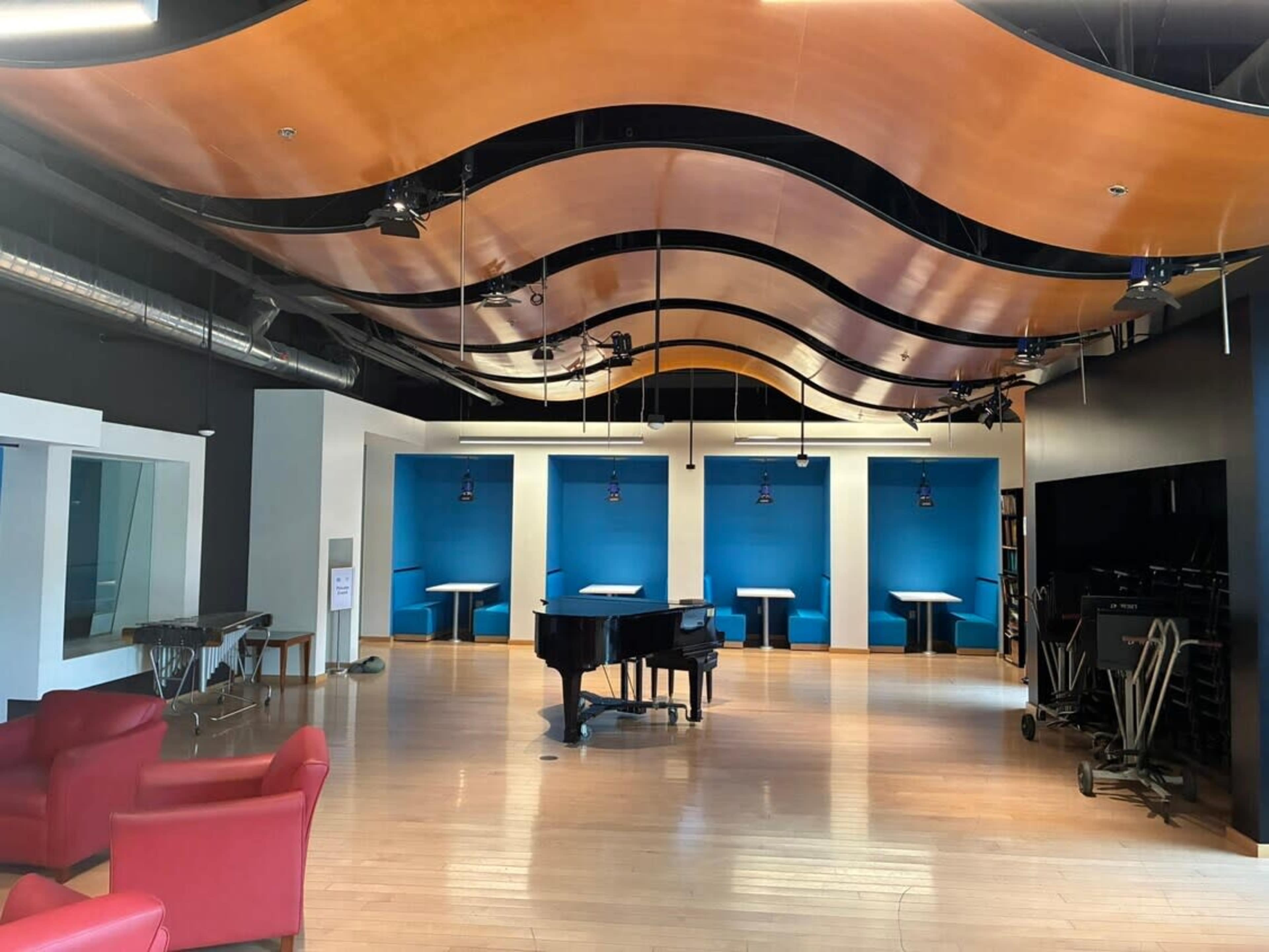 The image shows a modern, spacious room with a grand piano at the center, surrounded by blue wall panels and red seating.