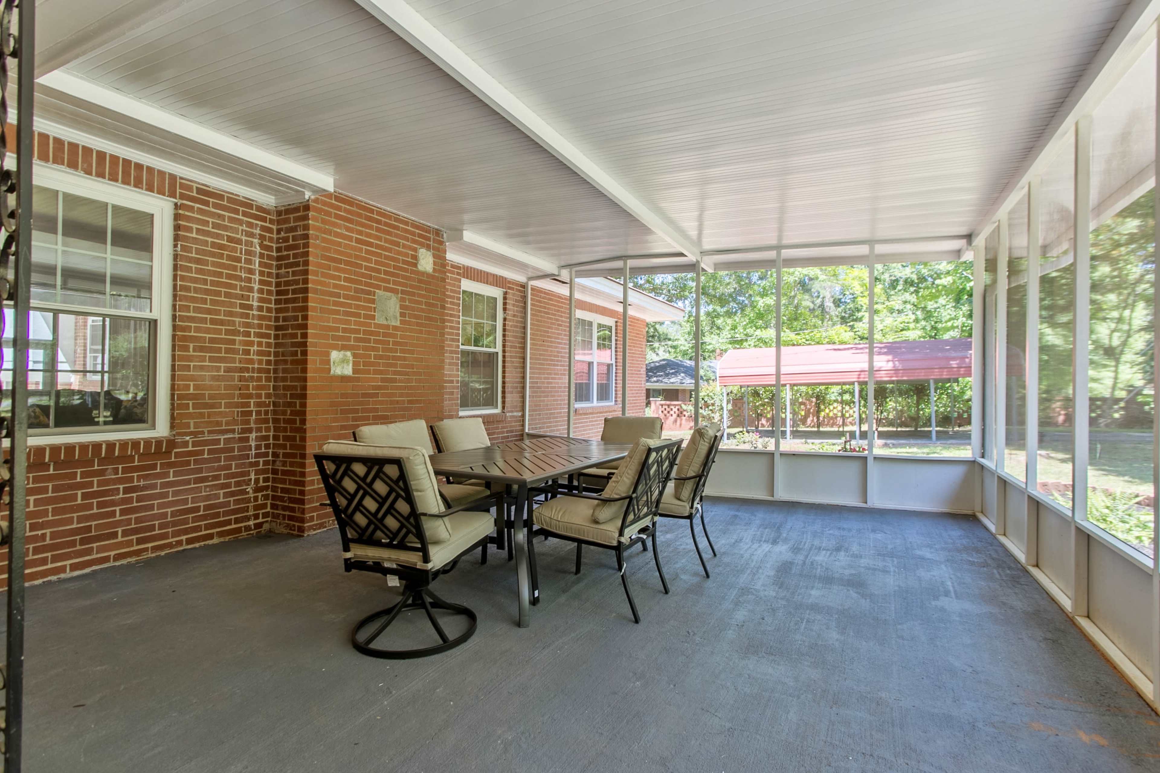 A screened-in porch featuring a rectangular dining table surrounded by six chairs on a concrete floor, with brick walls and large windows allowing views of the outdoors.