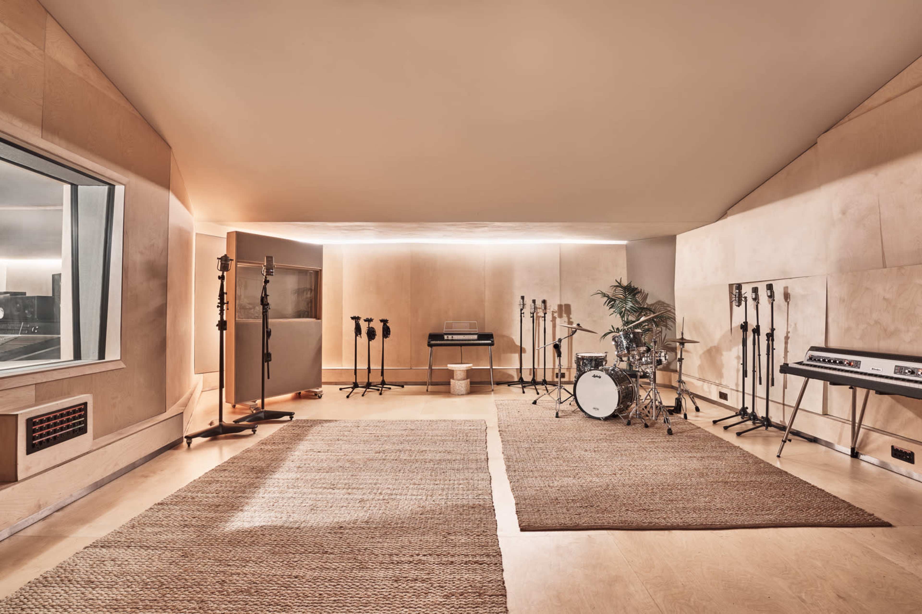 A spacious music studio features a drum set, keyboards, and multiple microphones arranged on stands, with a large window and natural light illuminating the room.