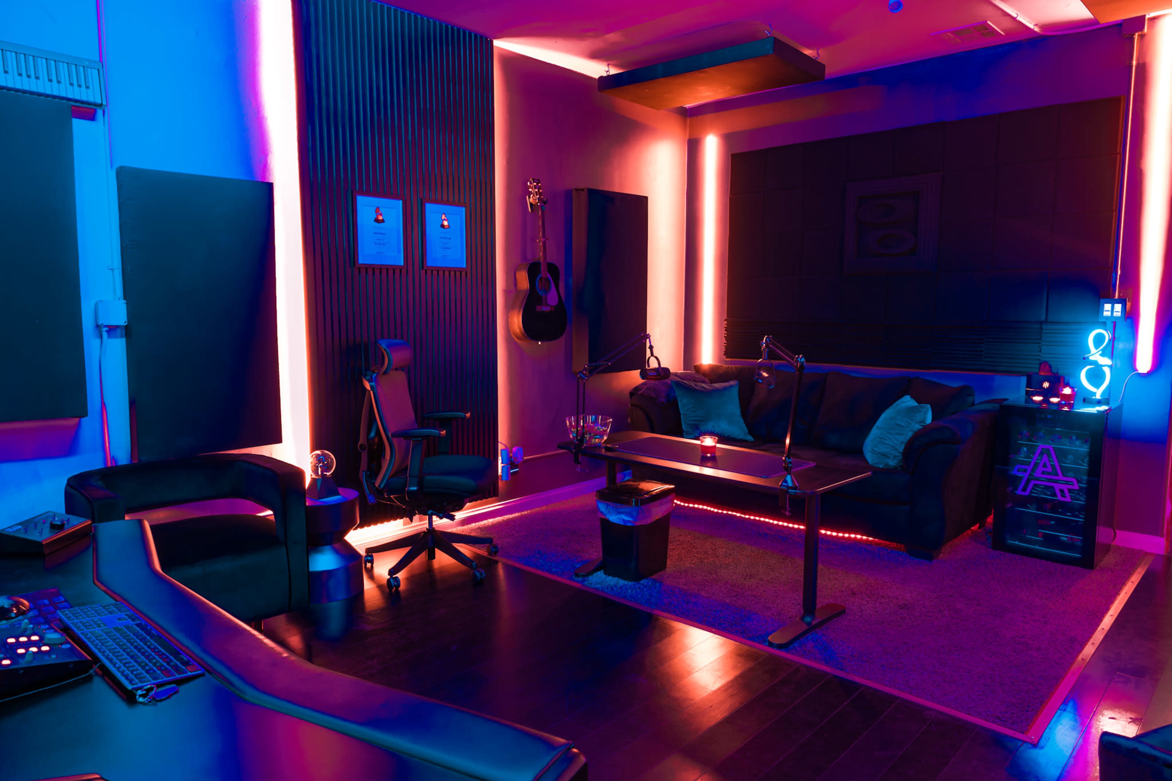 The image shows a modern recording studio with vibrant colored lighting, a couch, a guitar hanging on the wall, and a control desk with audio equipment.
