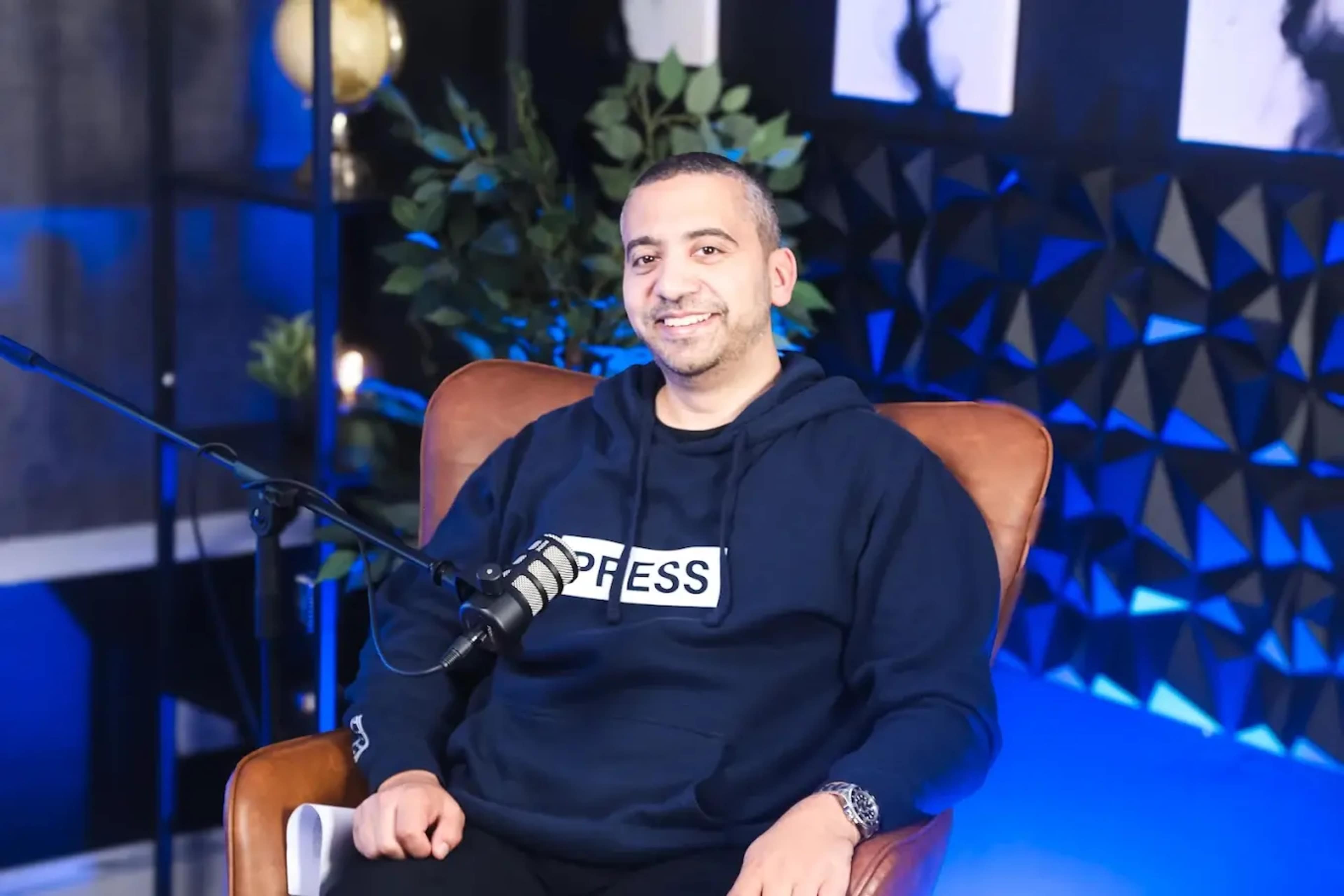 A man in a navy hoodie sits in a leather chair in a studio setting with blue lighting and decorative plants.