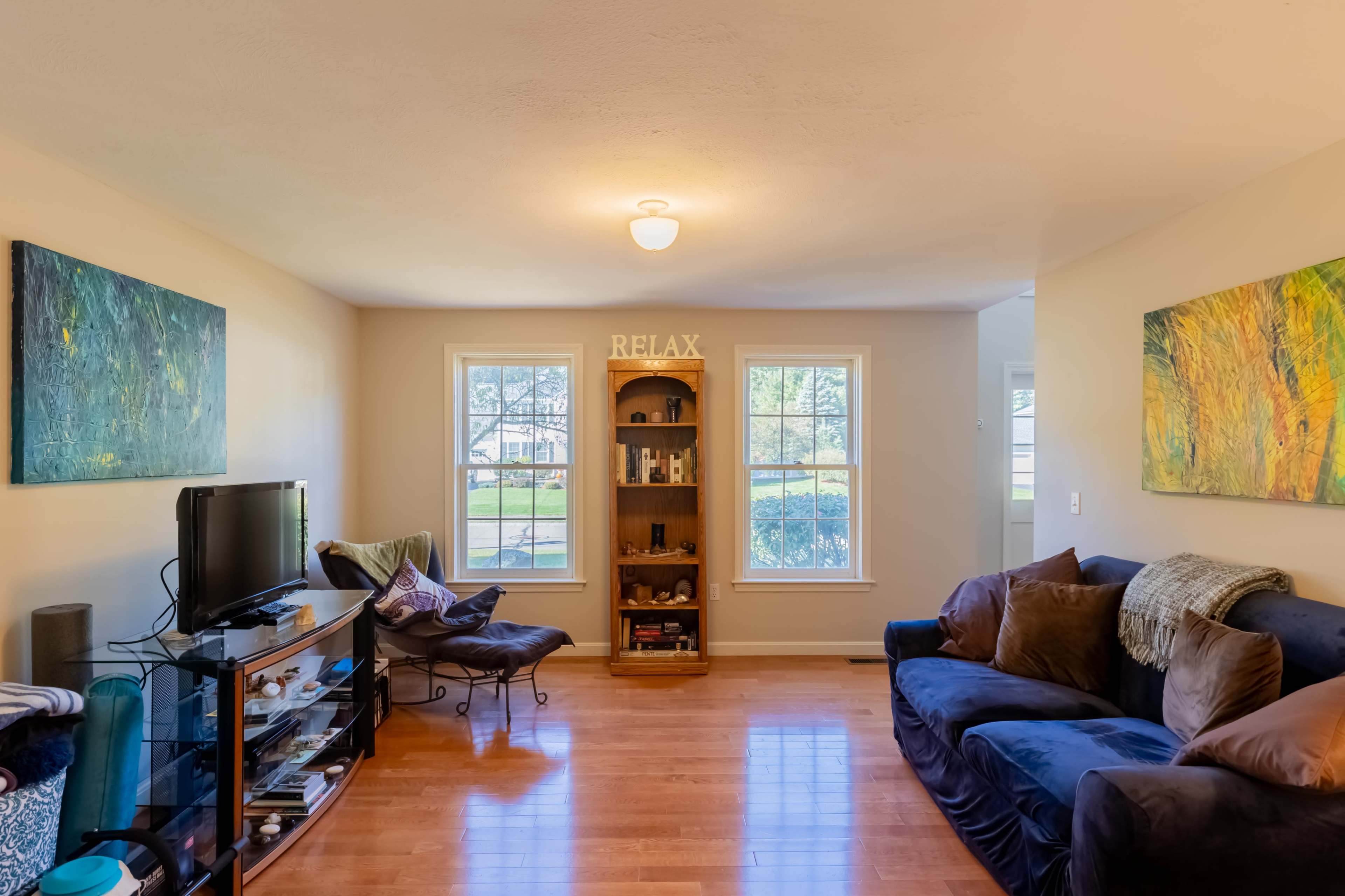A living room features a dark sofa, a glass television stand, and two large windows with artwork on the walls.