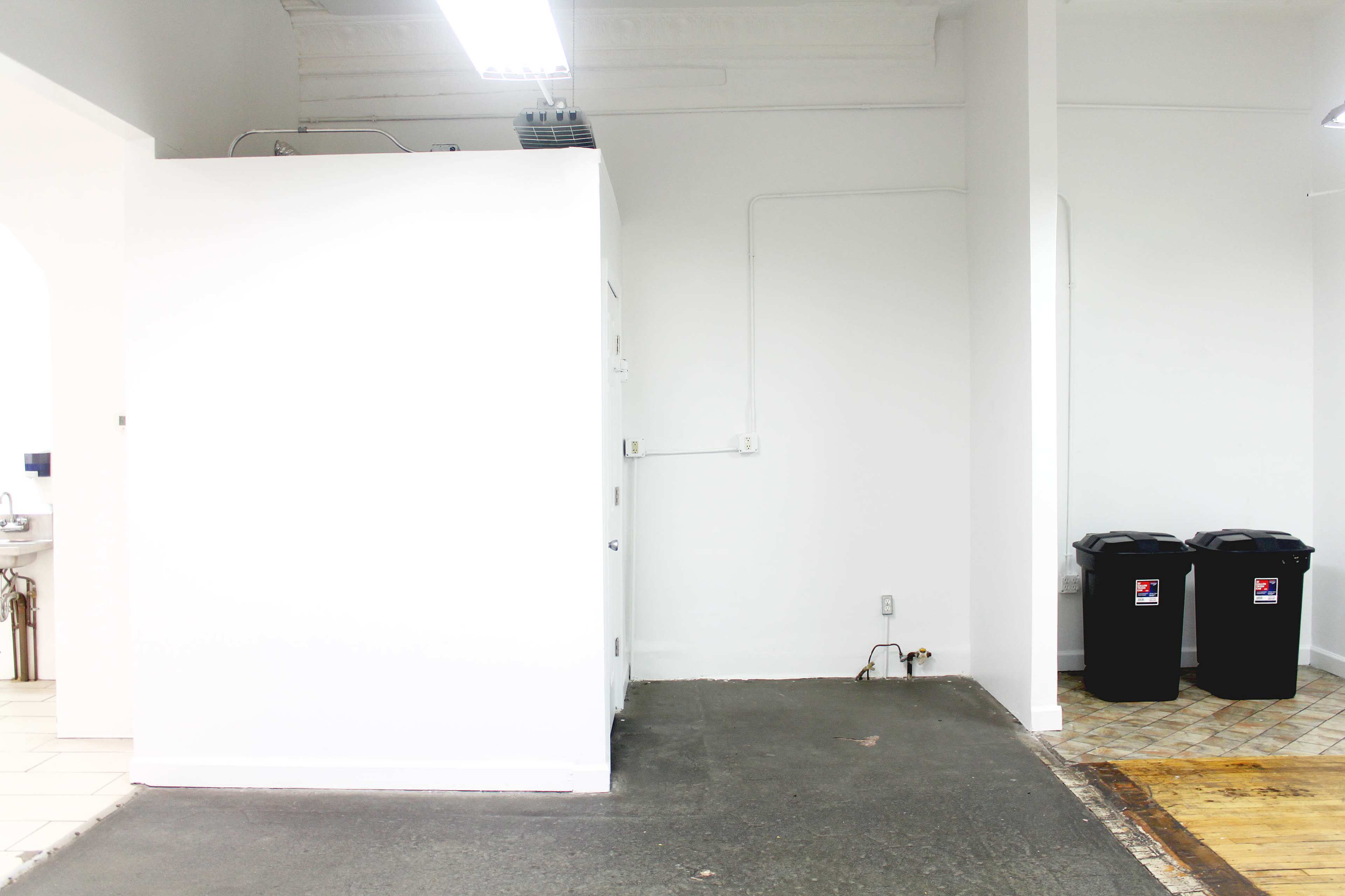 The image shows a bright, empty room with a white wall and two black trash cans on the floor by the wall.