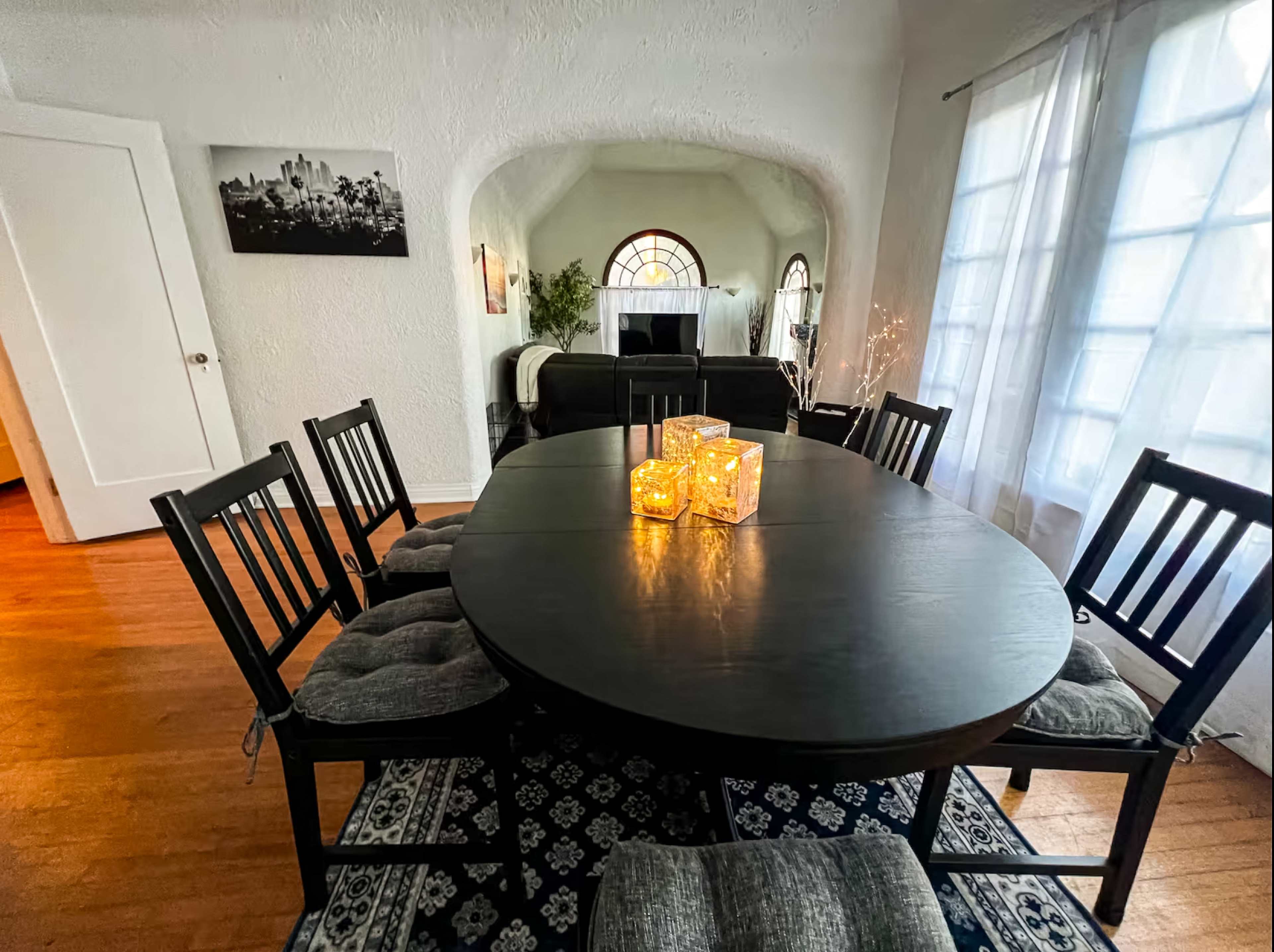 A dark wooden oval dining table is set with three illuminated candles, surrounded by six chairs, in a light-filled room featuring an arched doorway and large windows.