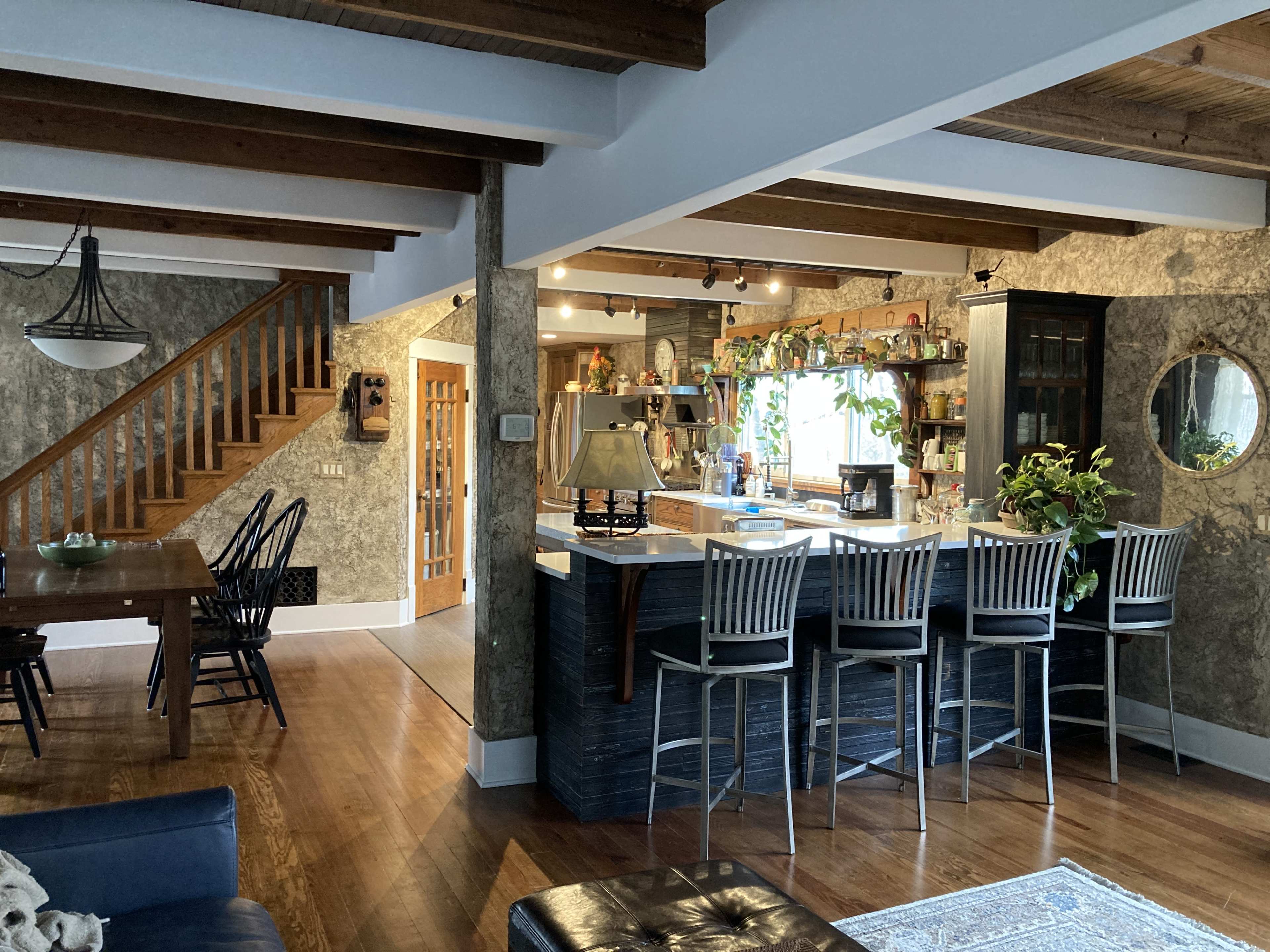A spacious kitchen and dining area features wooden beams, a central island with bar stools, and a staircase leading to an upper level.