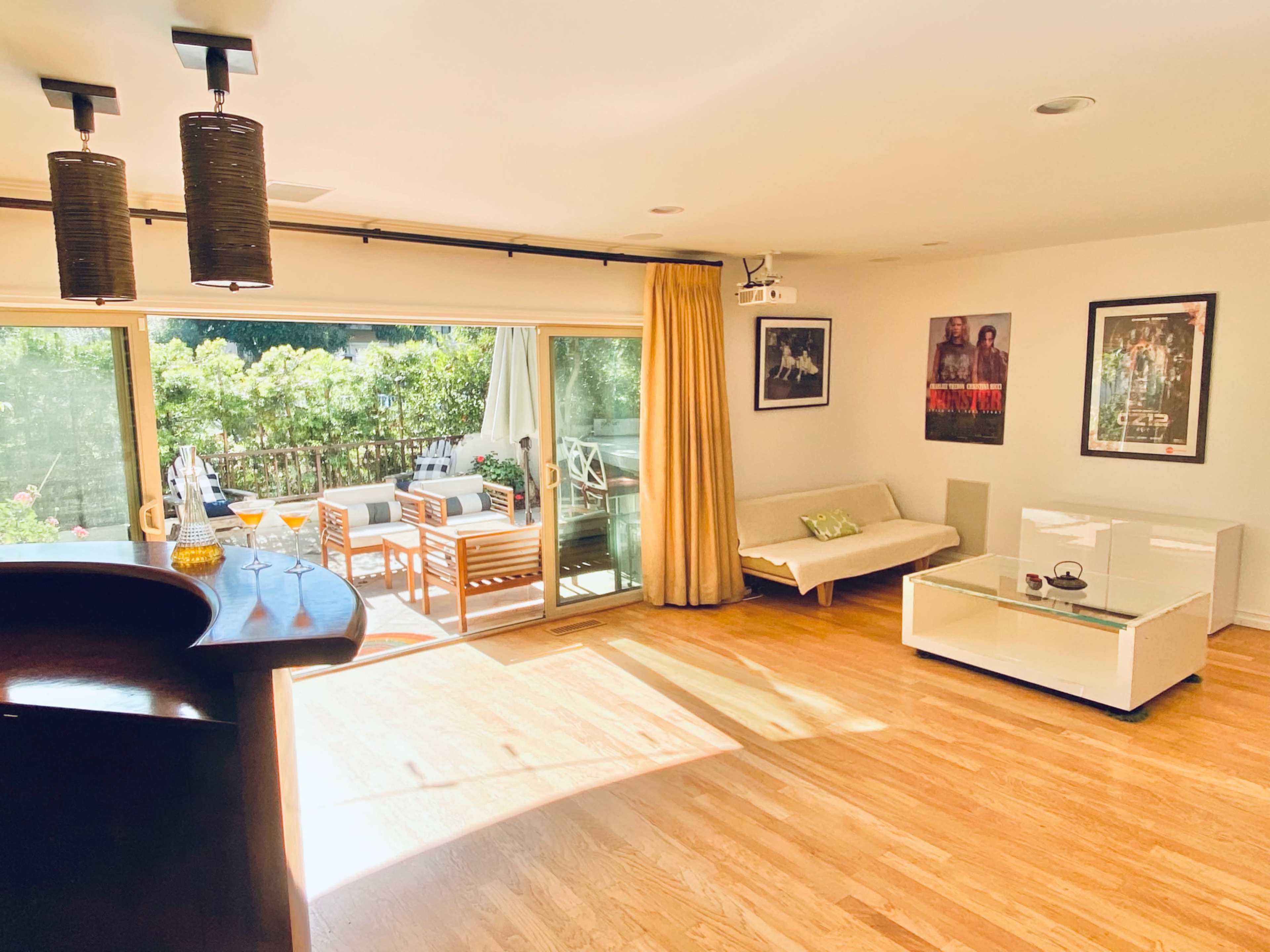 A bright living room with wooden flooring, a bar area, a sofa, and a sliding glass door leading to an outdoor seating area.