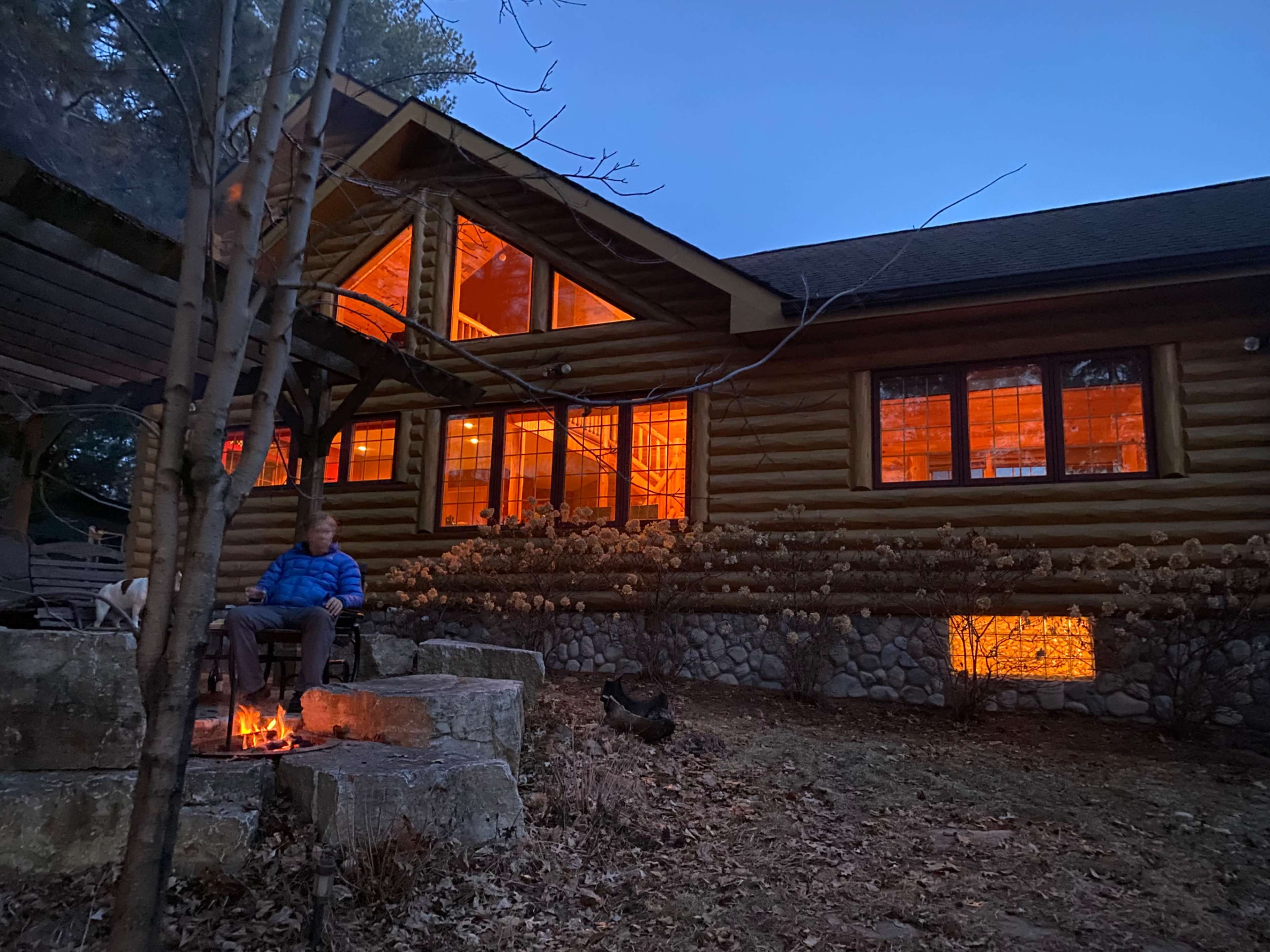 A person sits by a fire in front of a log cabin with illuminated windows as dusk approaches.