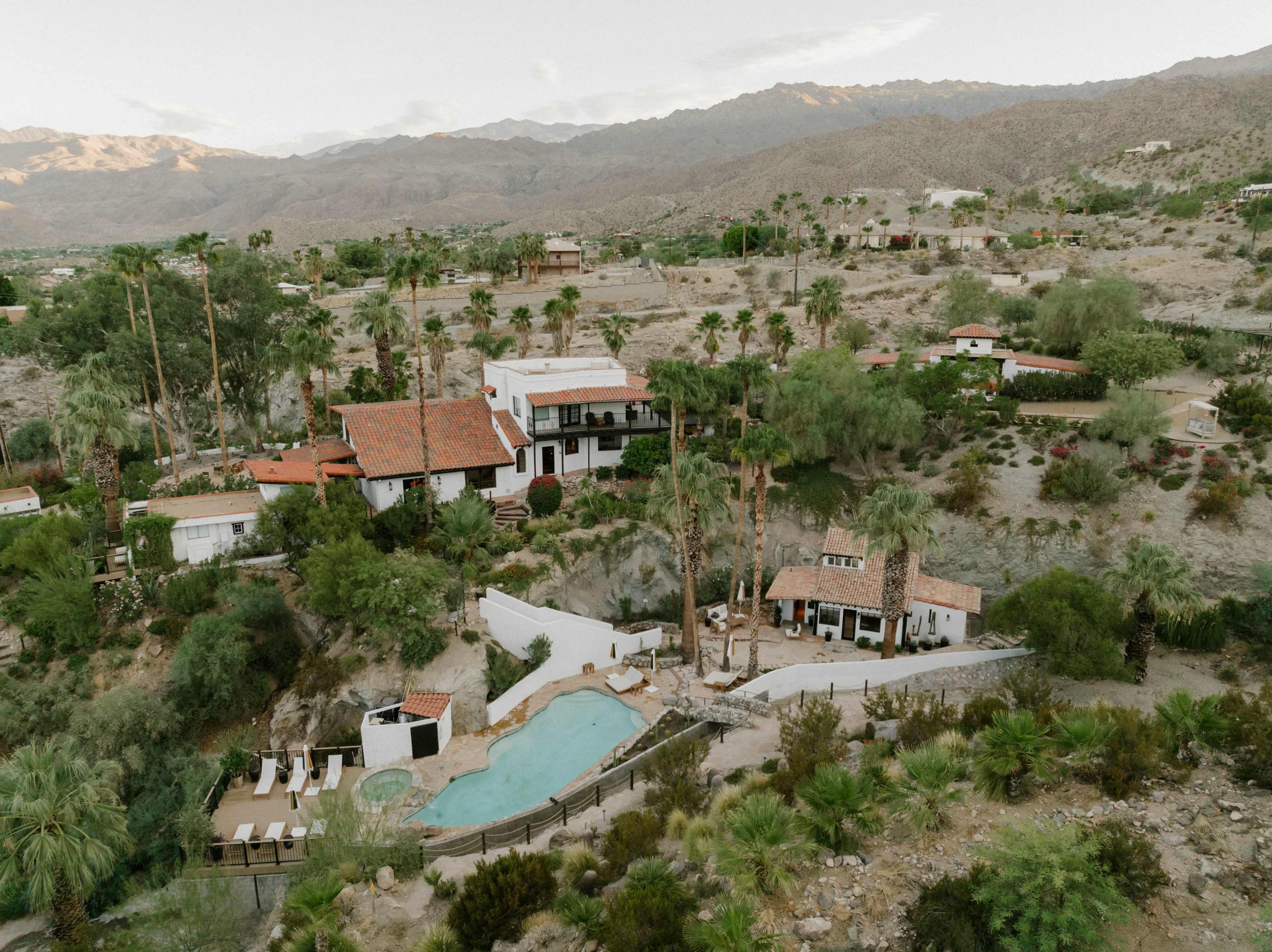 The image shows a hillside estate with several buildings, a swimming pool, and palm trees, surrounded by mountains in the background.