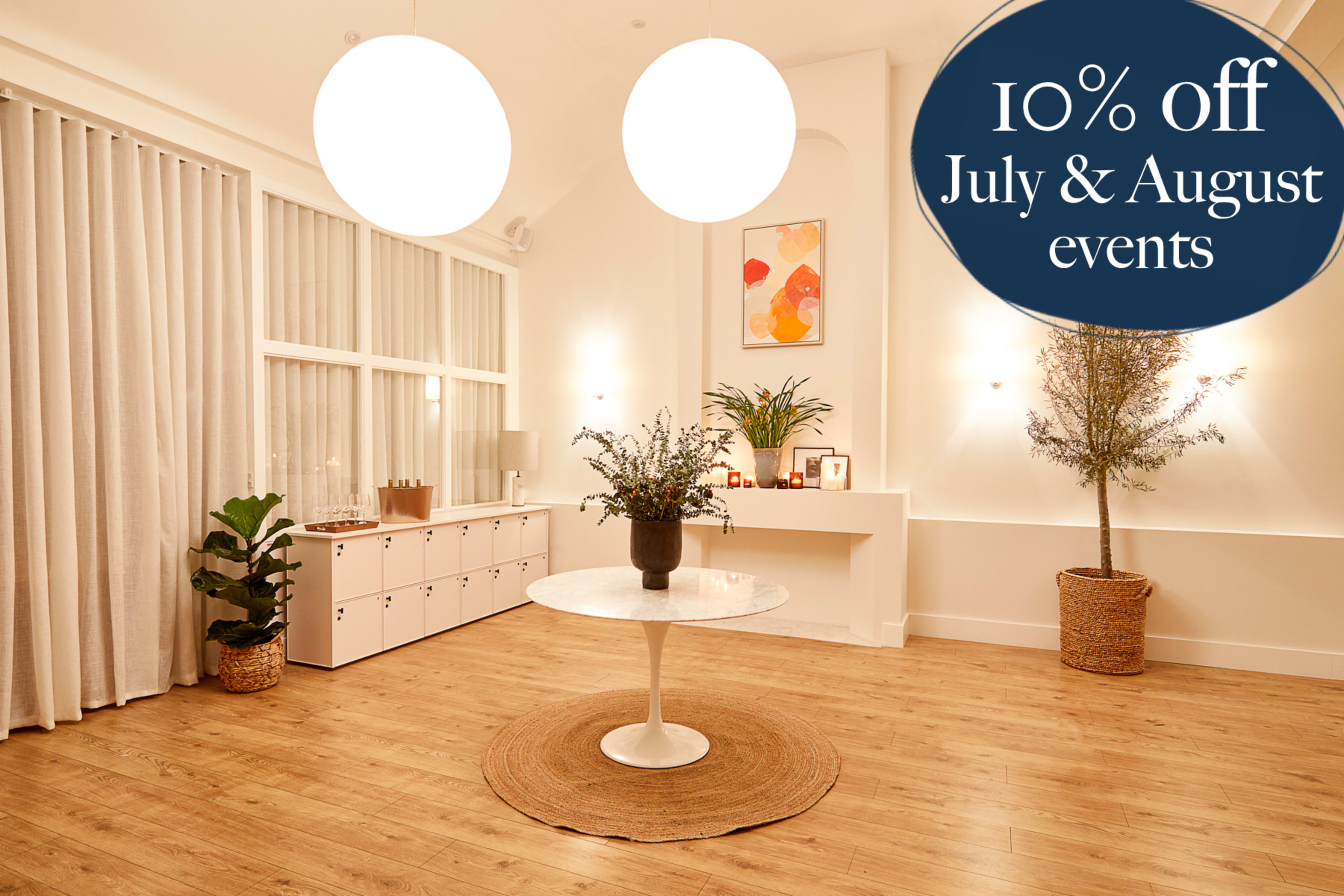 The image shows an elegantly decorated interior space featuring a round white table, decorative plants, and large pendant lights.