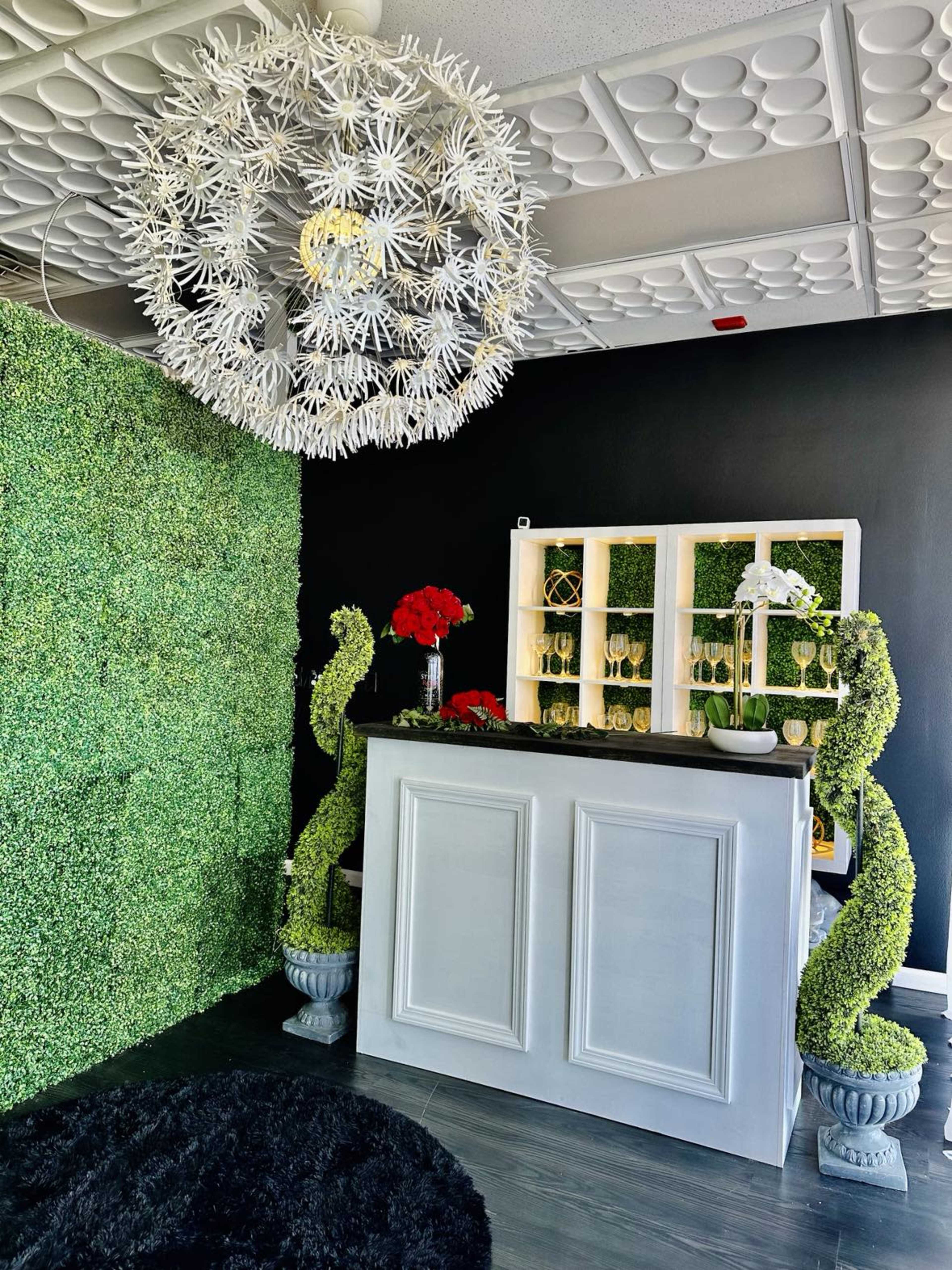 A reception area featuring a white counter, a green hedge wall, and decorative elements including a large chandelier and floral arrangements.