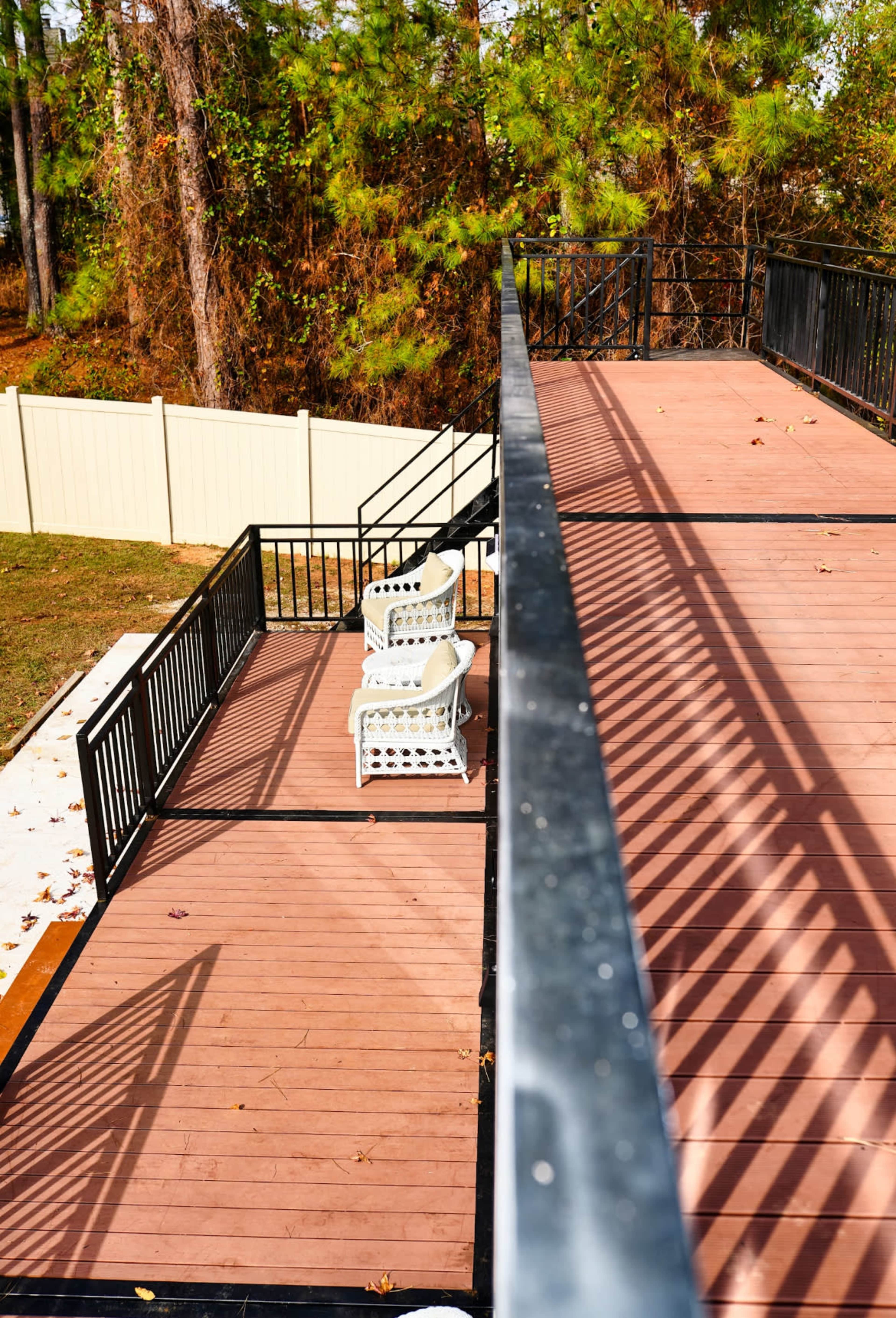 A second-story deck features two white chairs surrounded by a wooden surface and a railing with shadows cast by the sunlight.
